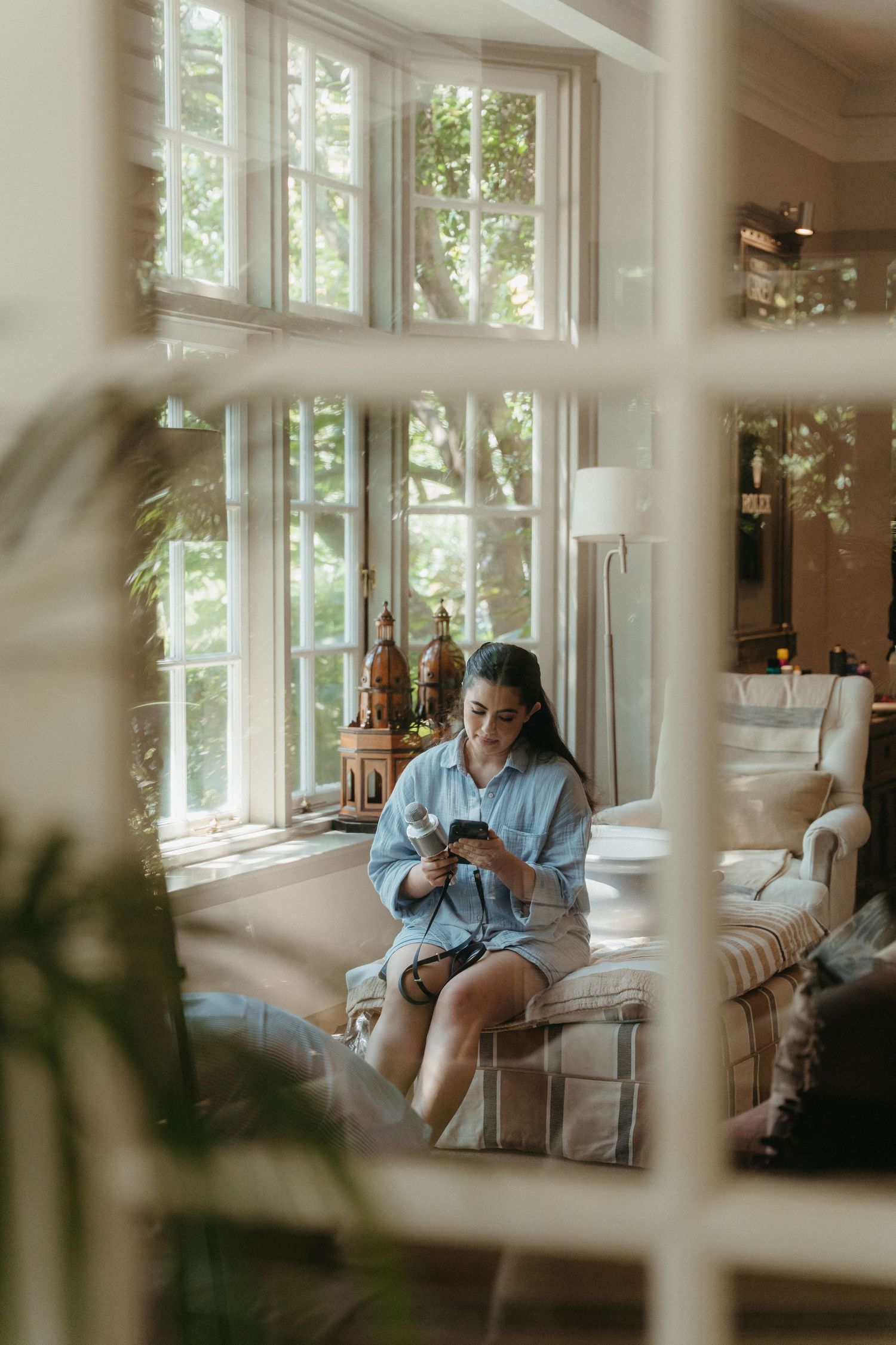Woman in blue shirt sits in armchair using phone in bright living room with large windows and green trees outside.