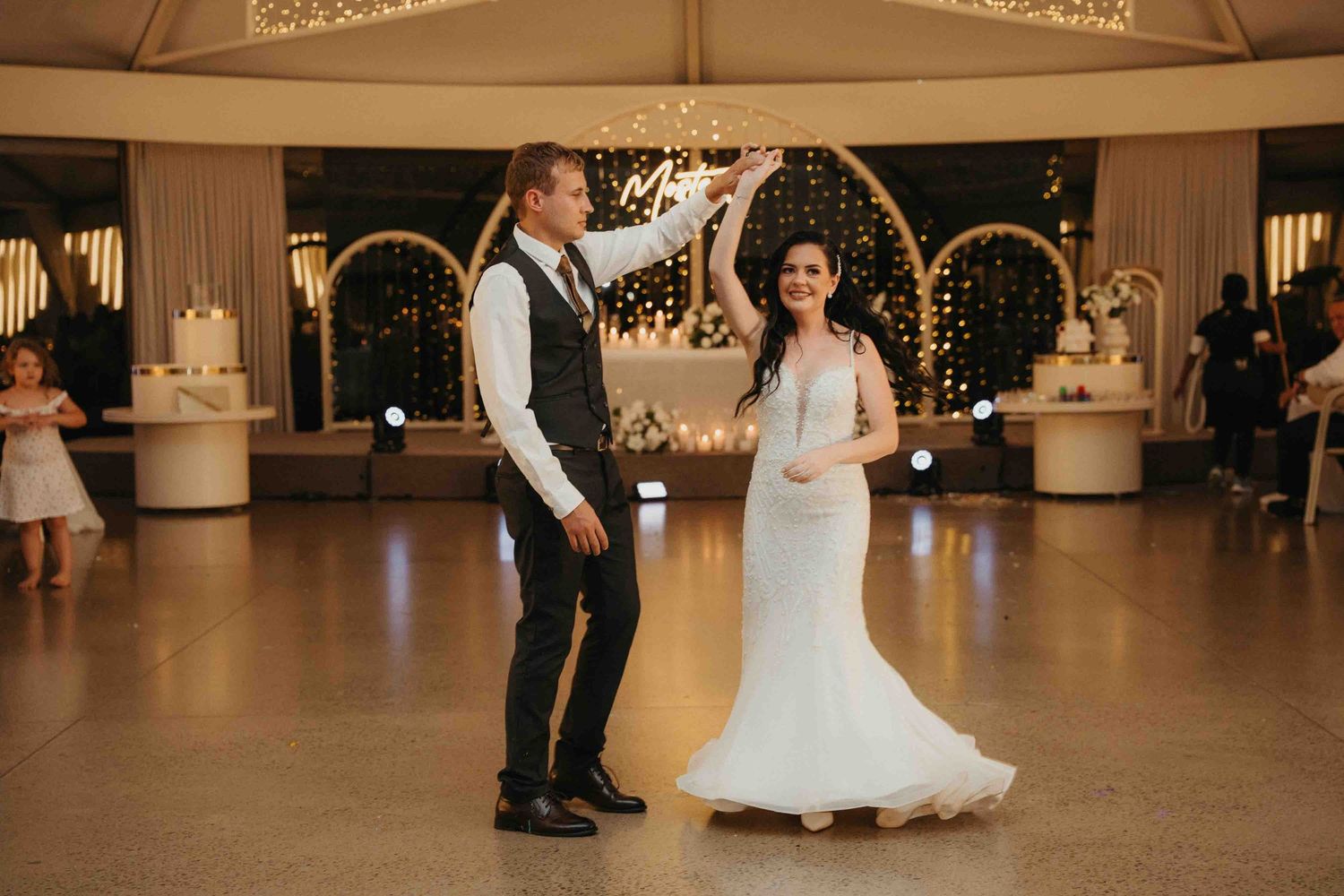 Bride and groom share first dance on elegant white dance floor with golden arch backdrop and warm fairy lights.