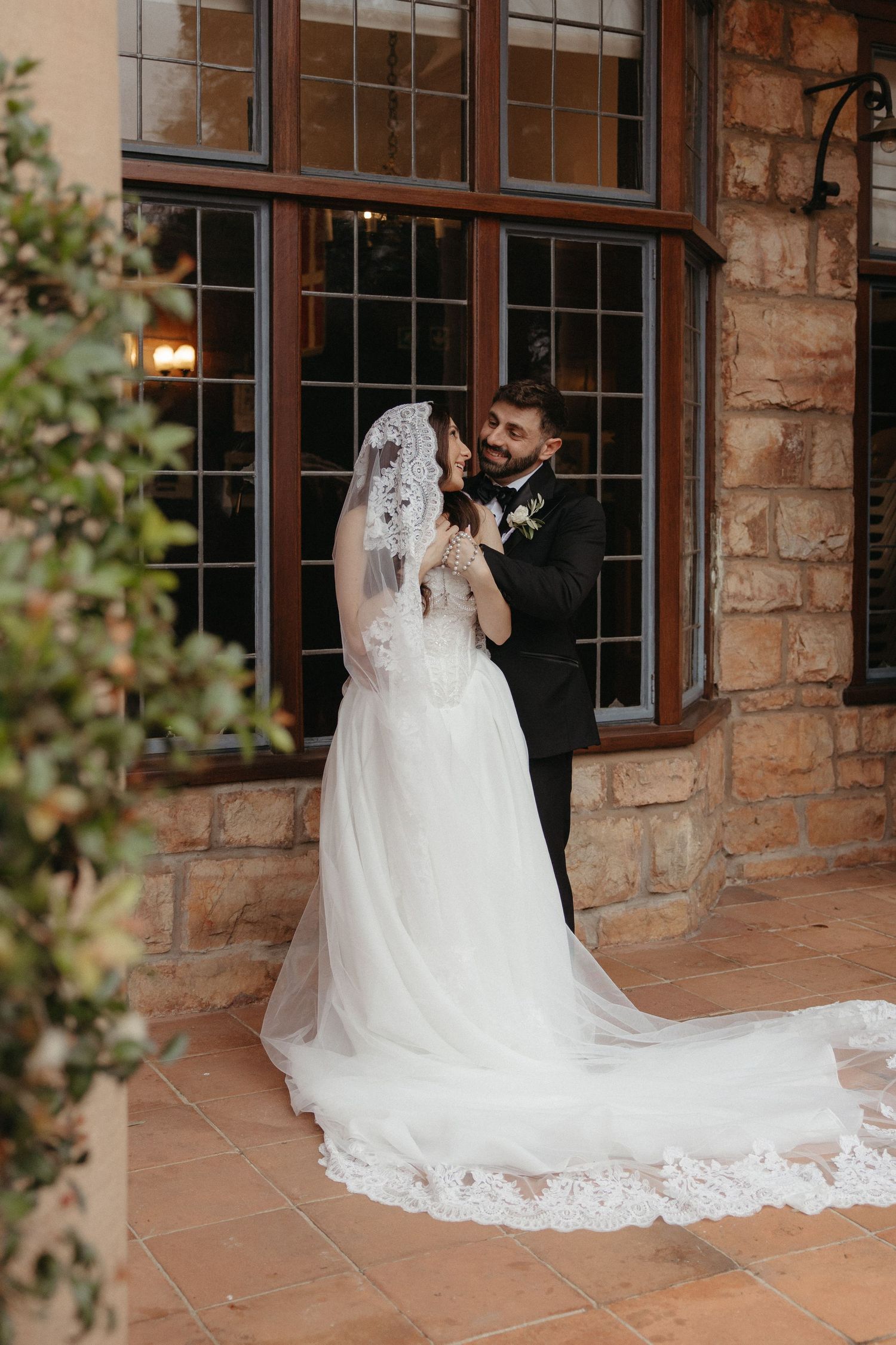 Bride in lace veil and groom in black tuxedo share a tender moment for wedding photos at Glenshiel