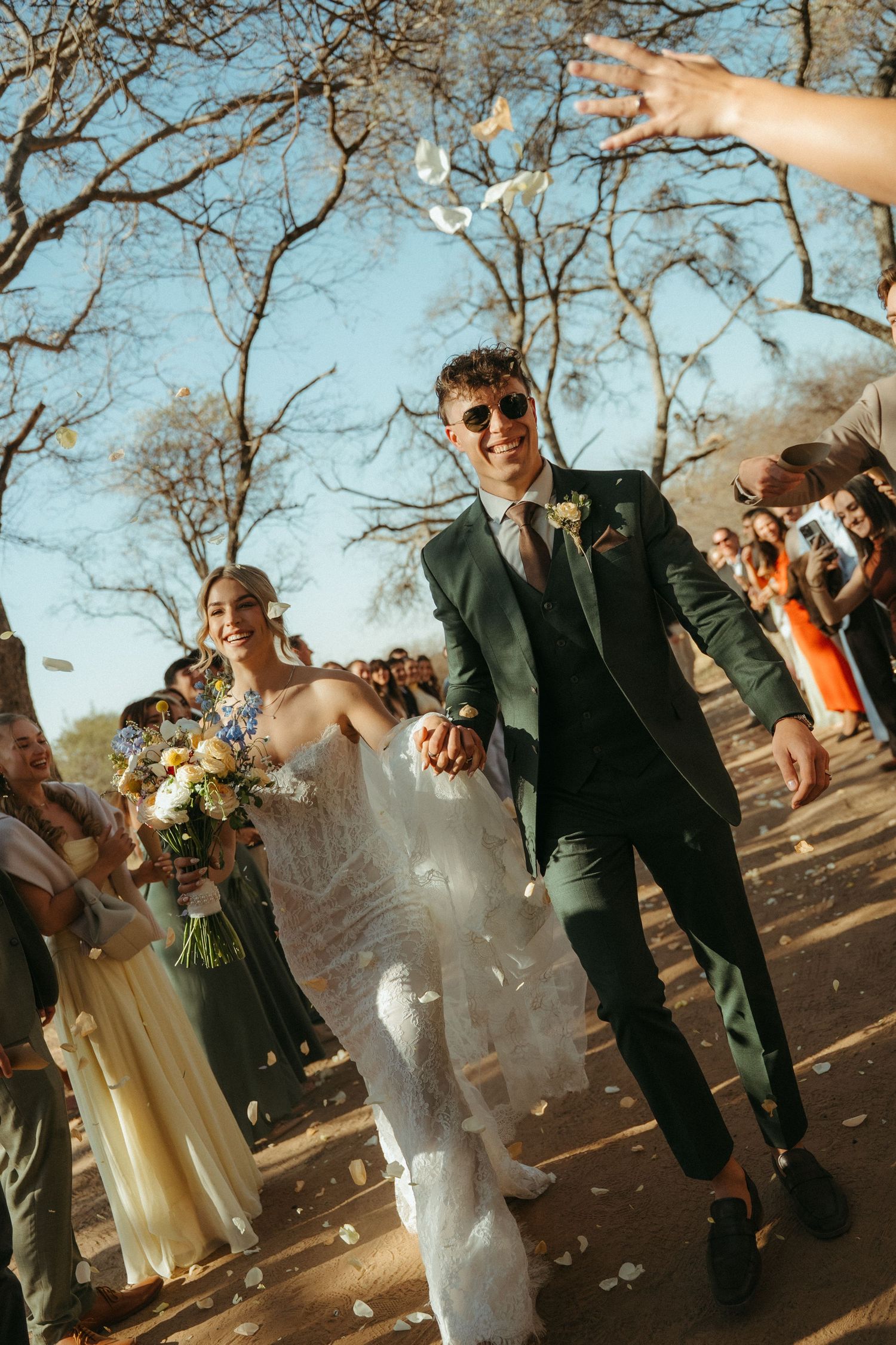 Joyful newlyweds walk through confetti shower, groom in green suit with sunglasses, bride in white gown holding bouquet at Monate Lodge.
