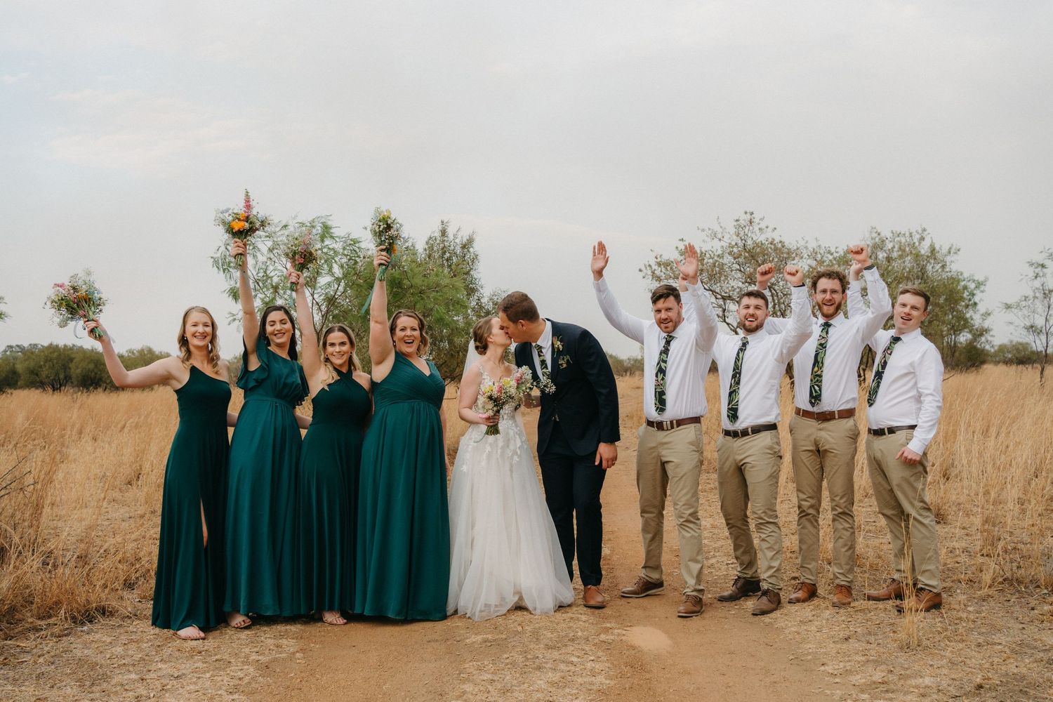 Wedding party in emerald dresses and khaki suits poses together in rustic outdoor setting.