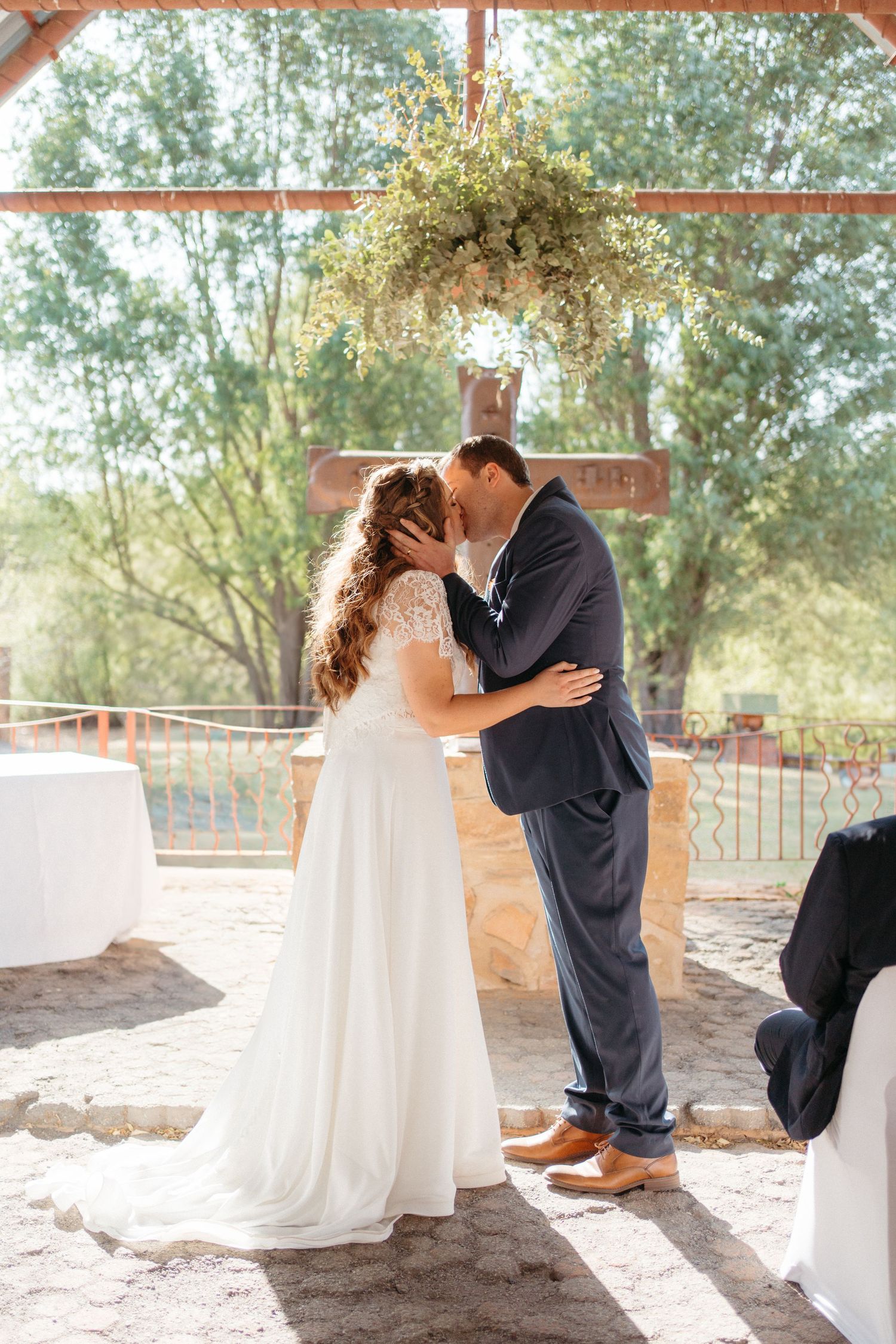 Romantic first kiss as newlyweds under floral archway at outdoor wedding venue.