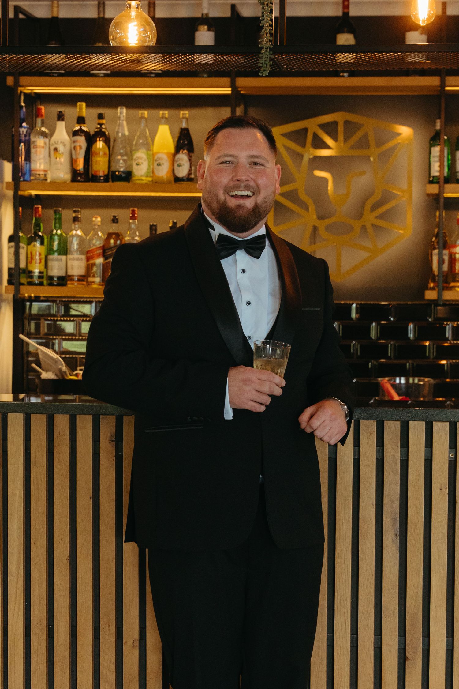 Professional bartender in tuxedo smiling behind an elegant bar counter with bottles and glassware displayed.