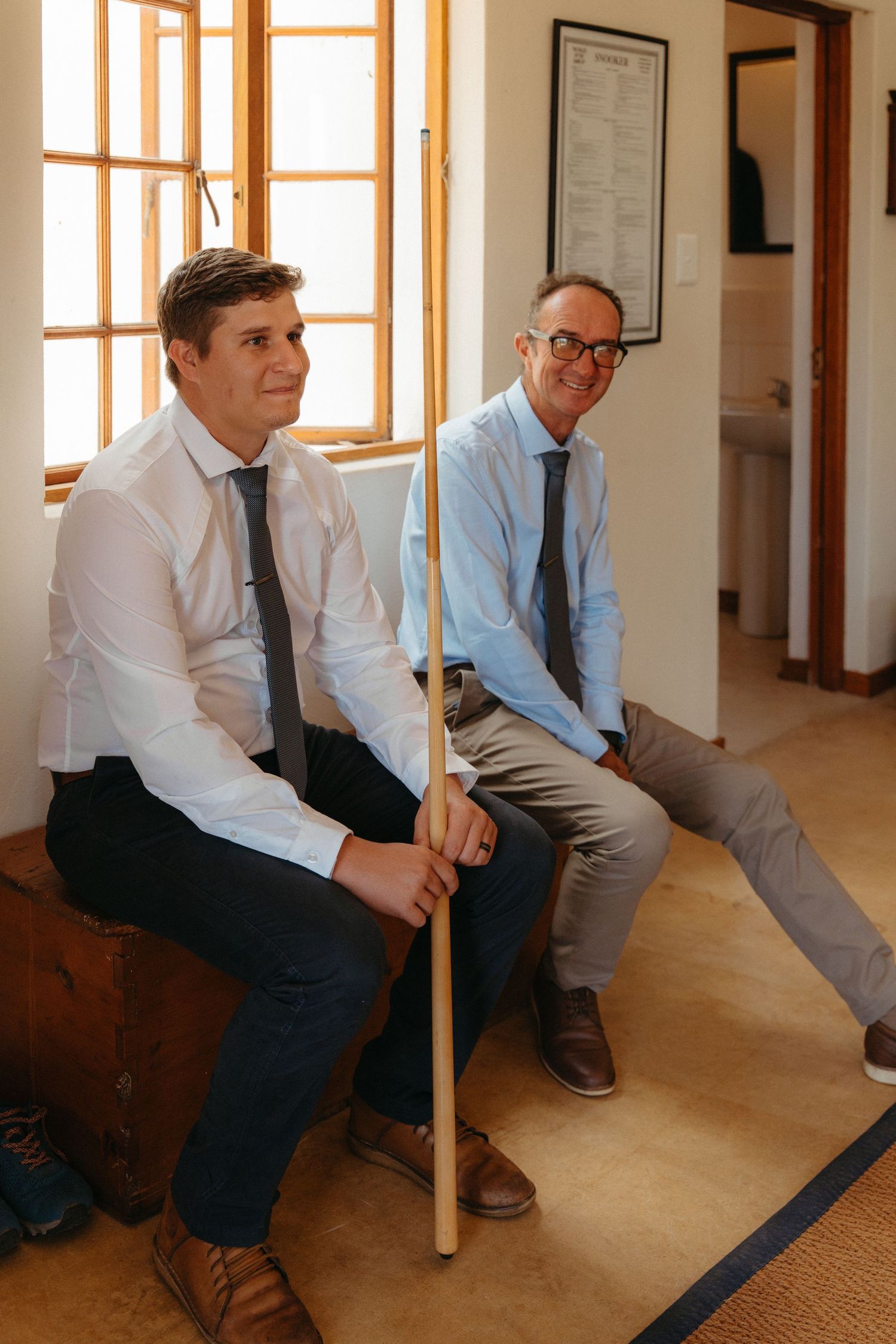 Two people in business attire sit on a wooden bench in a bright hallway with sunlit windows.