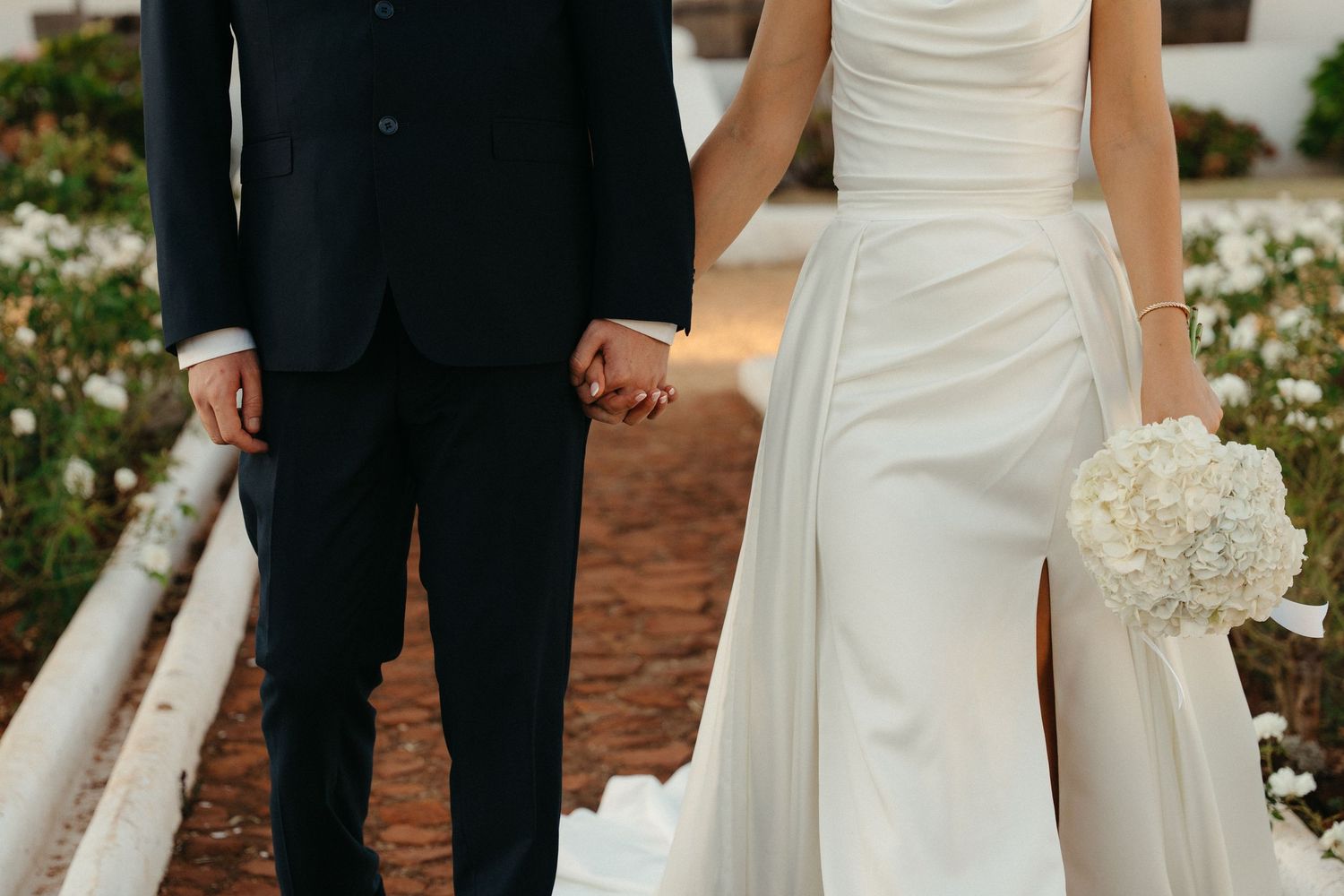 Wedding couple in formal attire holds hands while standing on brick path surrounded by white flowers and greenery.