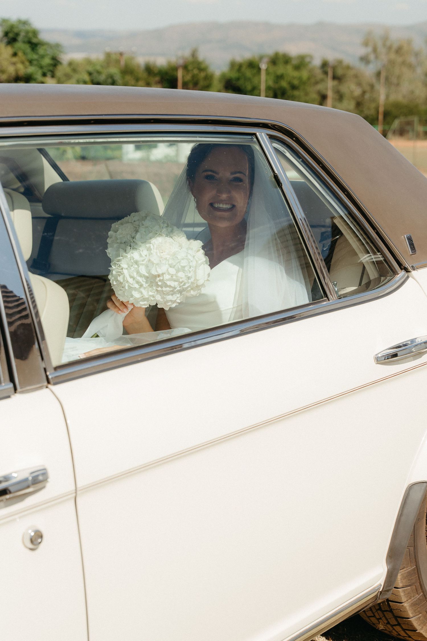 Wedding party riding in a classic white car with floral decorations through a scenic desert landscape.