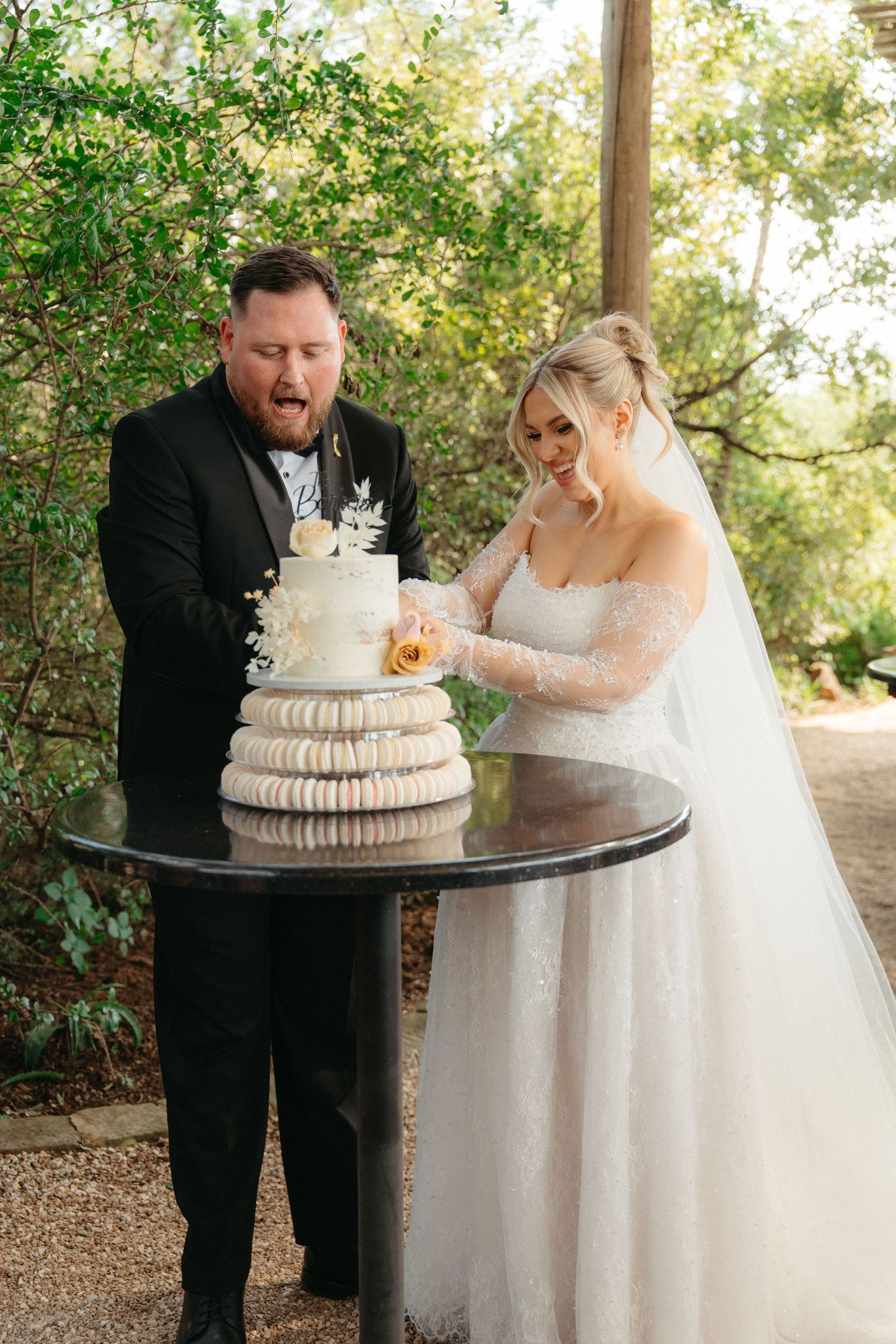 A couple cuts their elegant white tiered wedding cake together at an outdoor garden reception.