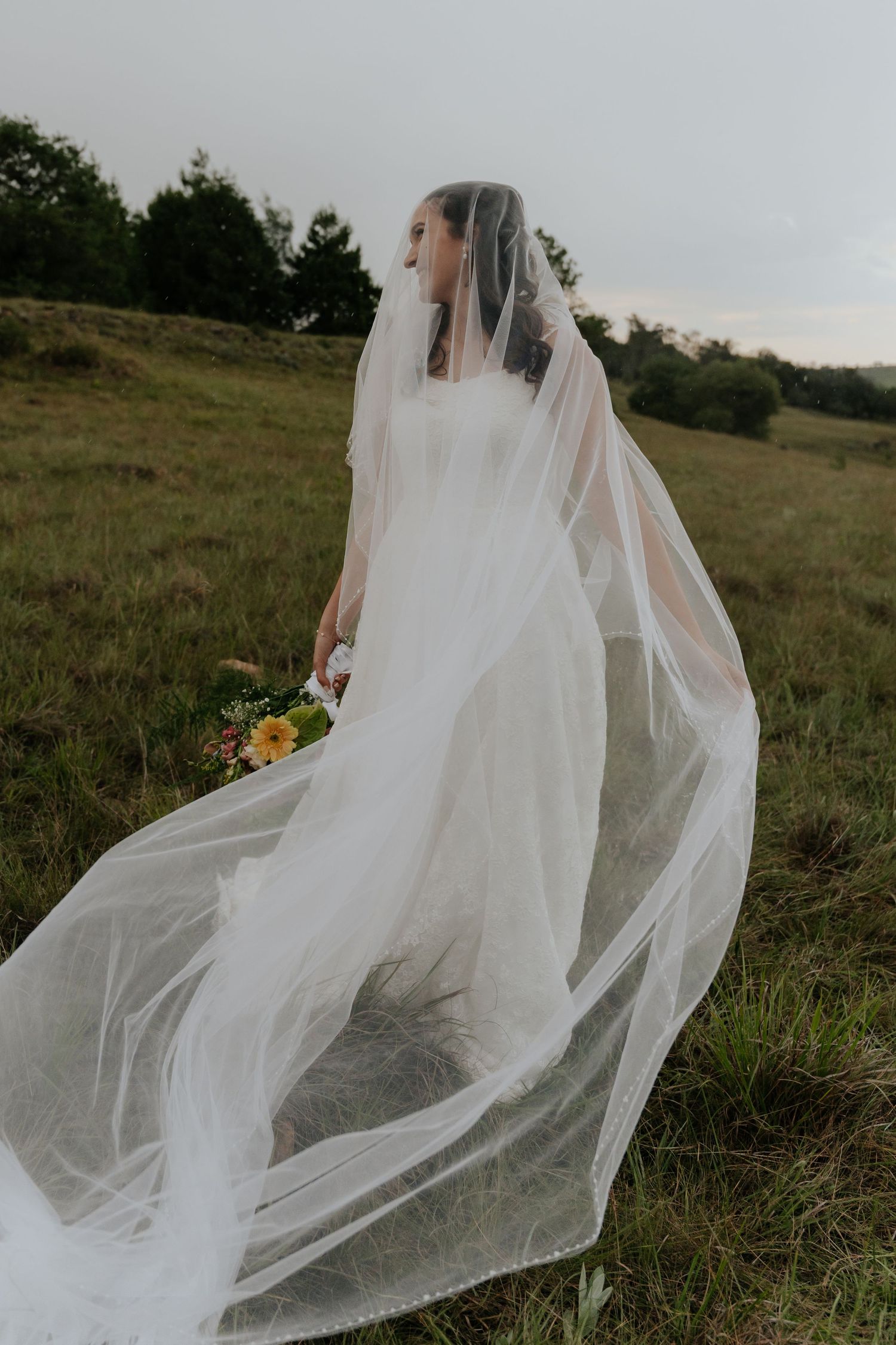 A flowing white wedding veil catches the breeze in a rustic outdoor field setting at sunset.
