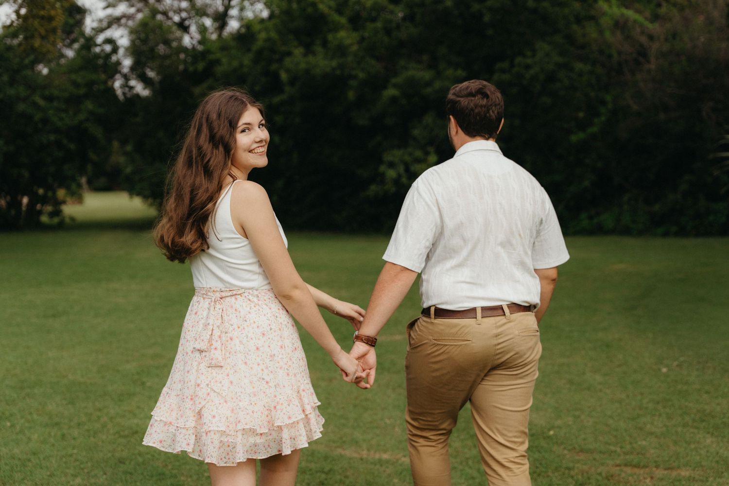 Couple holding hands in a green park, woman in floral skirt smiling over shoulder, man in white shirt and khaki pants.