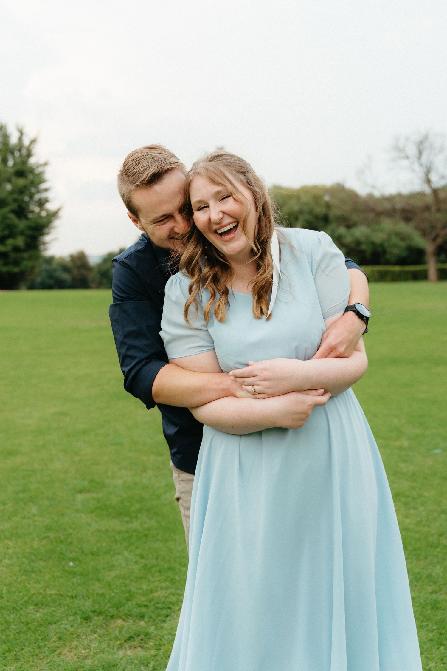 A couple shares a joyful embrace while standing in a green field on a cloudy day.