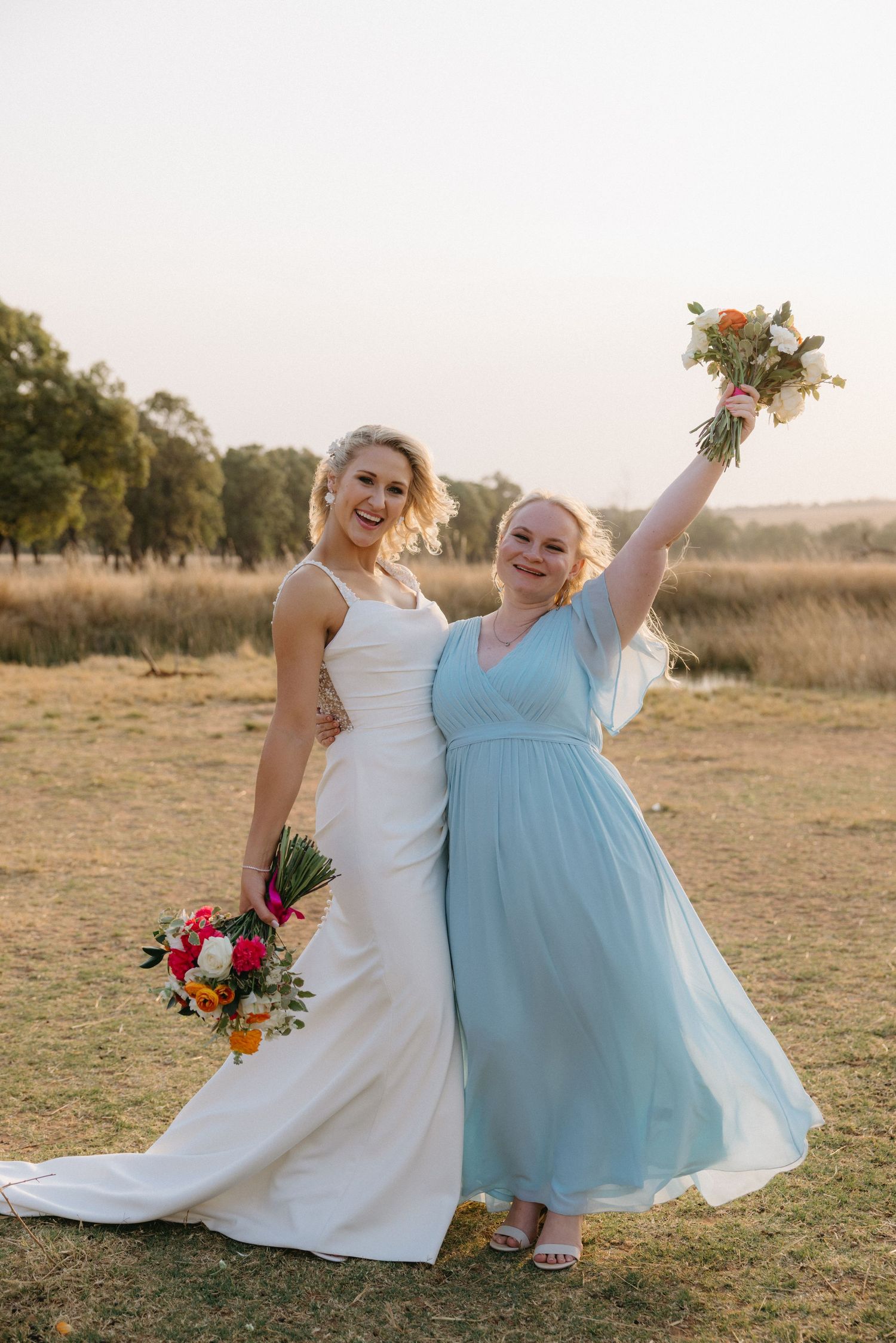 A bride in white and bridesmaid in light blue twirl joyfully together in a sunny outdoor field.