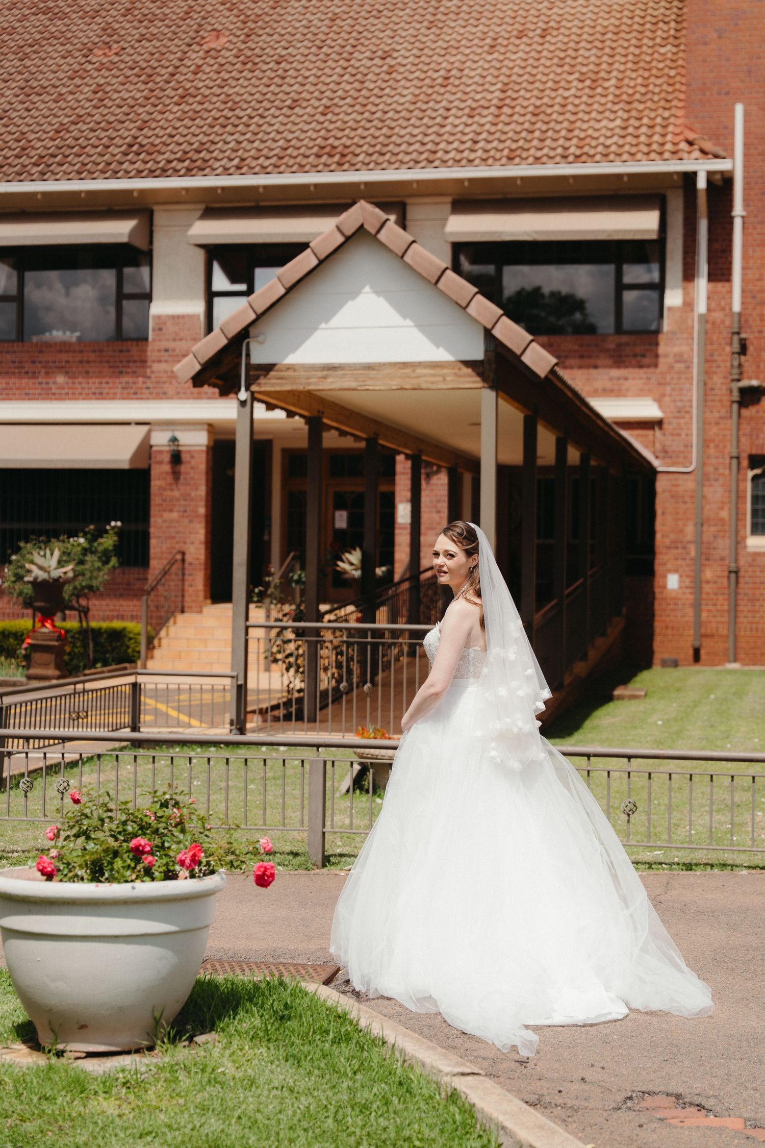 A bride in a flowing white ball gown and veil stands in front of a brick building with a covered walkway.