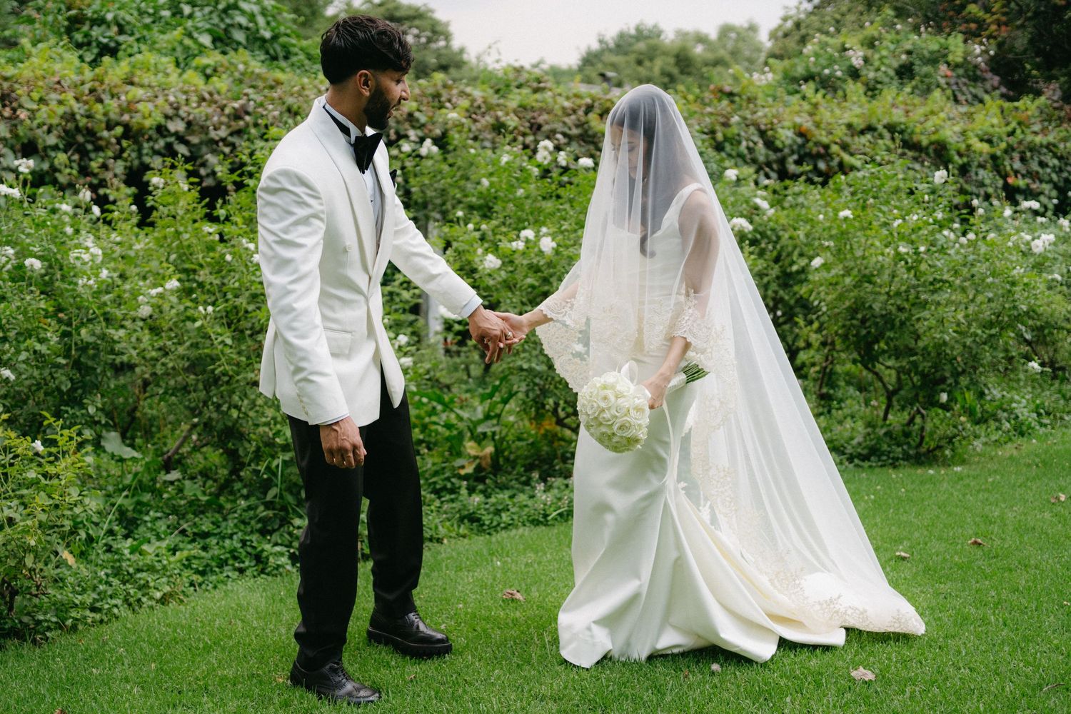 A couple celebrates their outdoor garden wedding, with the bride in white gown and veil holding hands with groom in a white jacket.