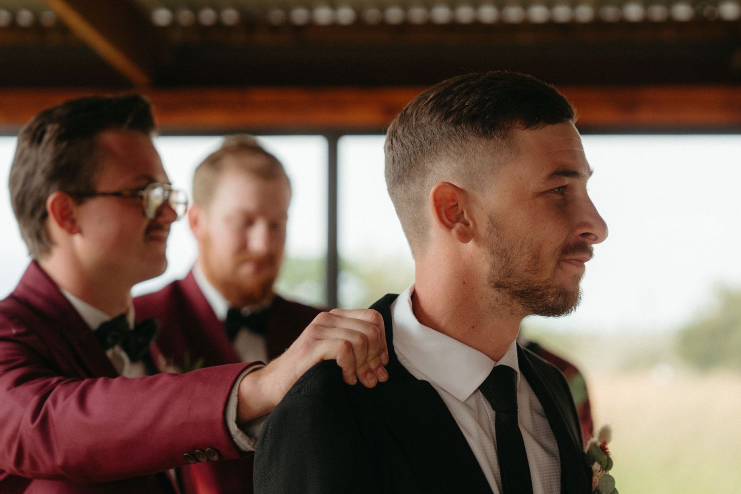 Groomsmen in burgundy attire helping with wedding preparations in a sunlit room.
