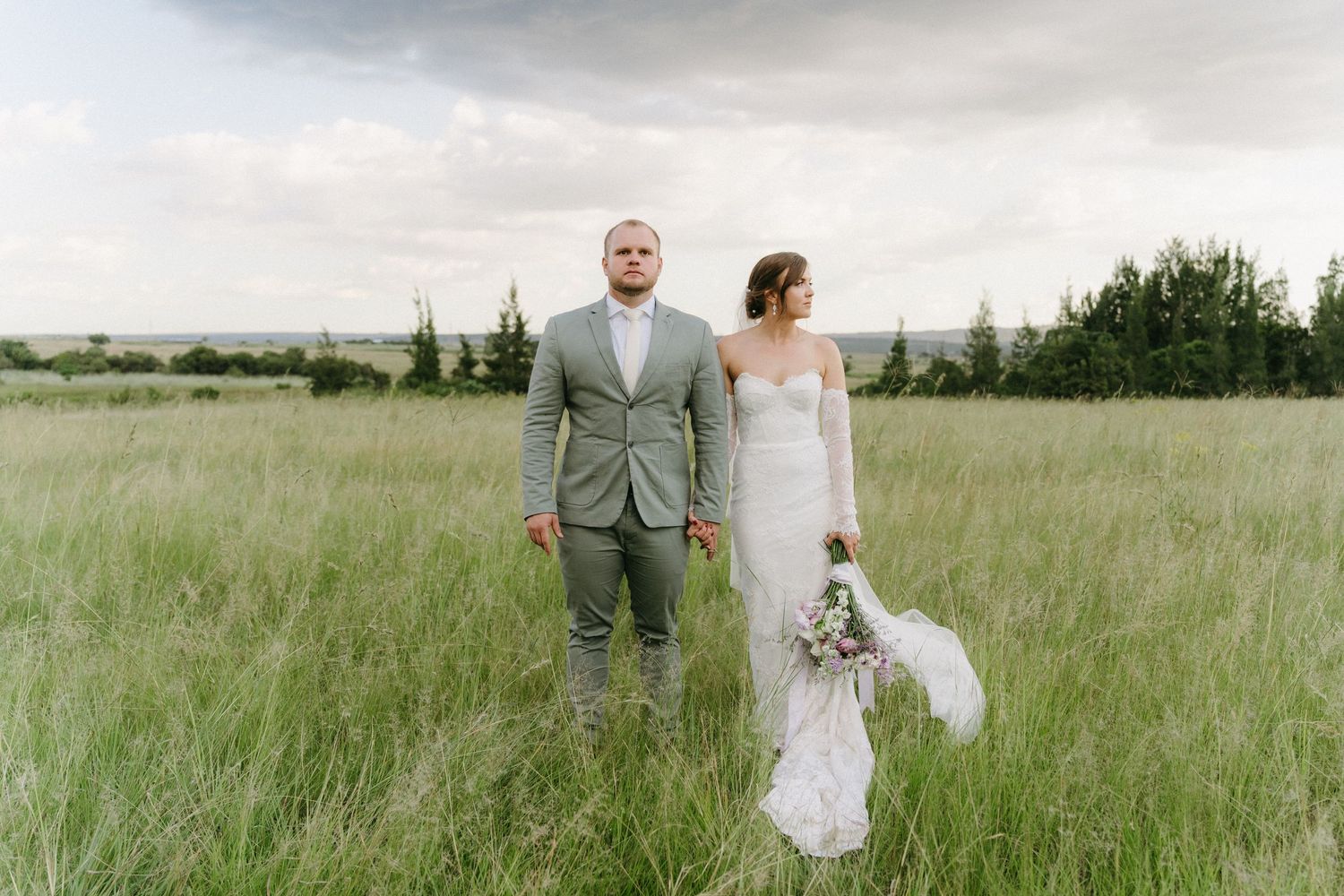 Newlywed couple stands together in a serene meadow under cloudy skies.