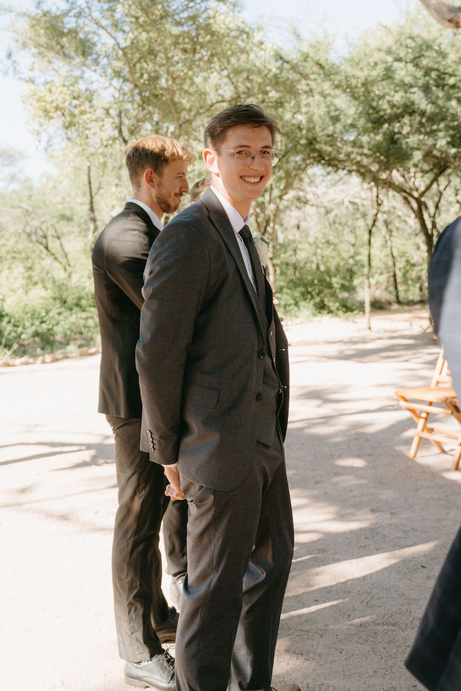 A formal wedding party member in a dark suit stands outdoors during a sunny daytime ceremony.
