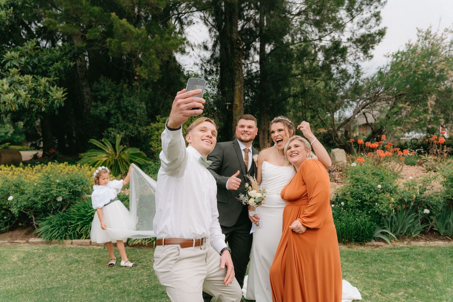 Wedding party in white and burnt orange dresses posing together on grassy lawn with trees.