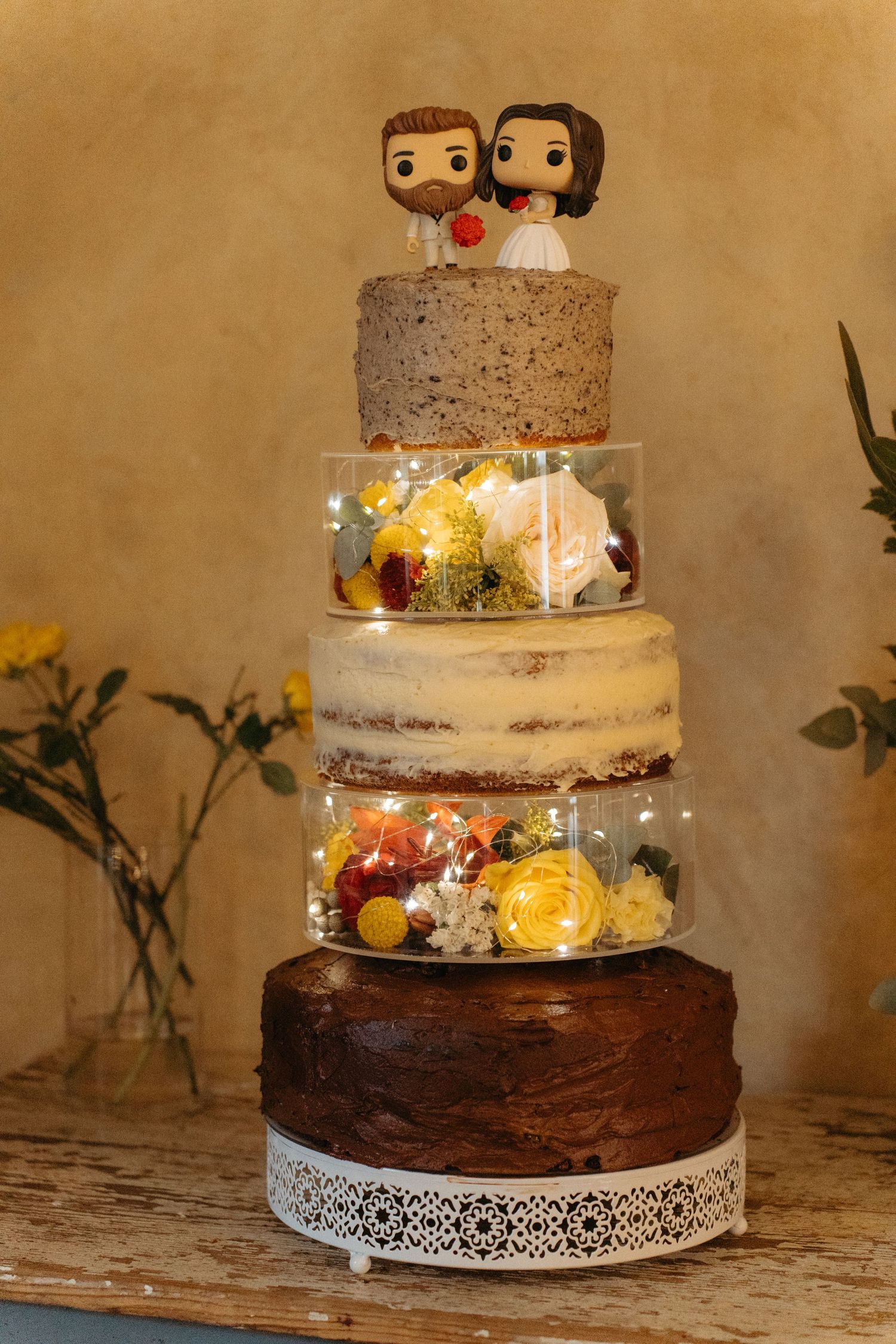 Rustic multi-tiered wedding cake with clear acrylic tiers filled with flowers and topped with cartoon bride and groom figurines.