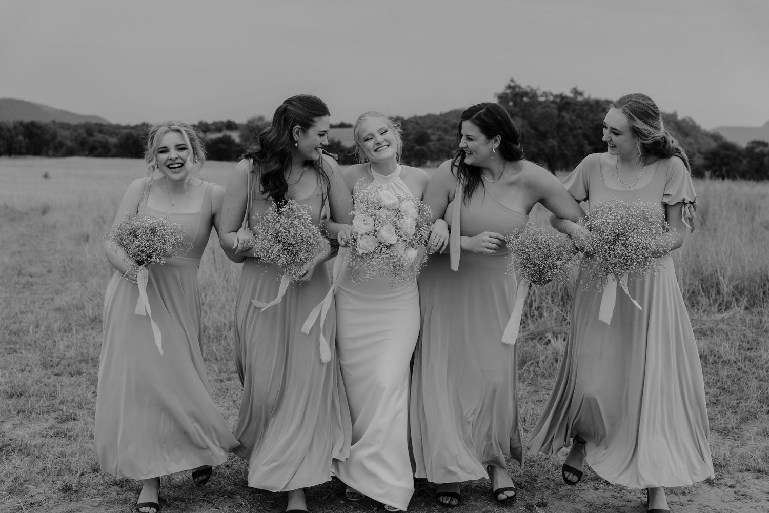 Black and white photo of wedding party sharing joyful moment with bouquets in rustic outdoor setting.