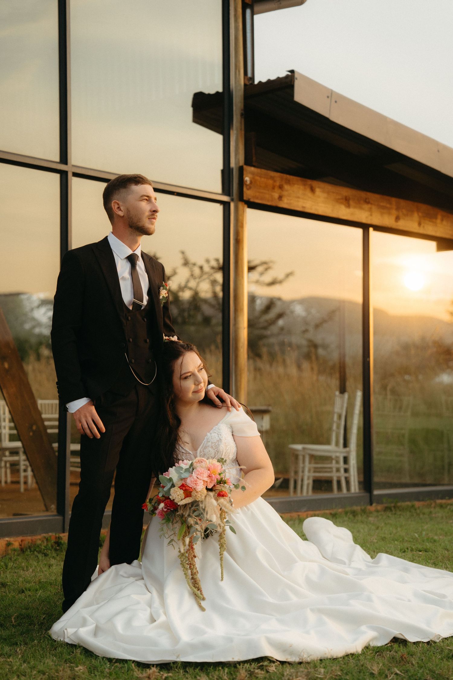 Elegant couple in wedding attire poses at sunset by large glass windows of modern venue with scenic outdoor view.