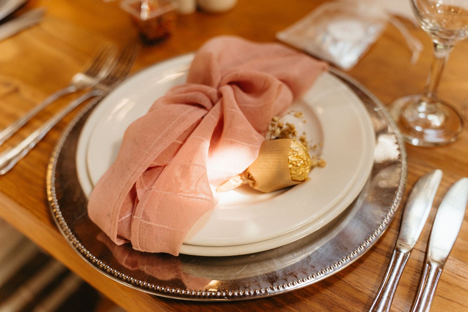 An elegant place setting with pink napkin, silver charger plate, and wine glasses arranged on a wooden dining table.