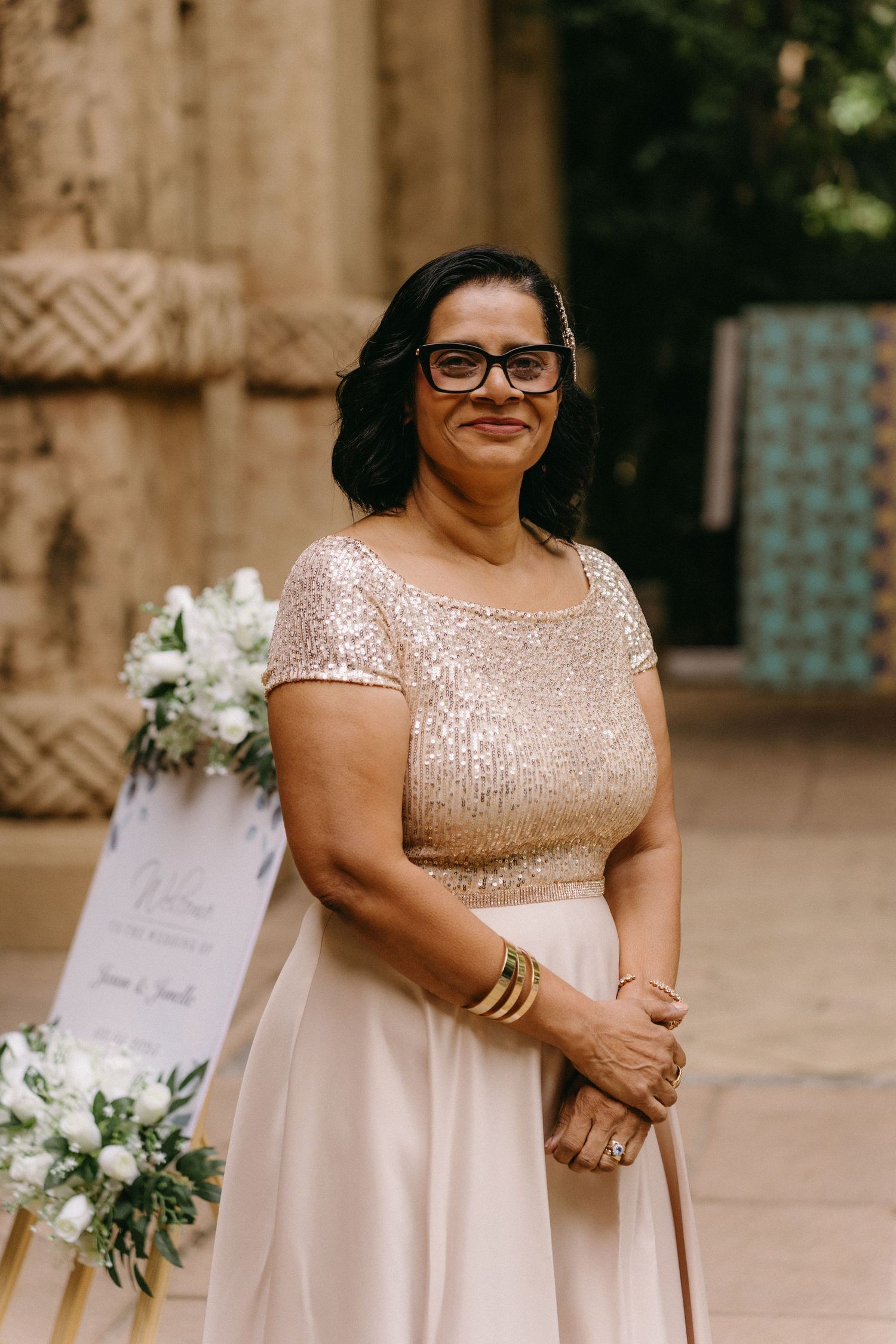 Wedding guest wearing a blush pink sequined dress and glasses stands elegantly at an outdoor venue with floral decorations.