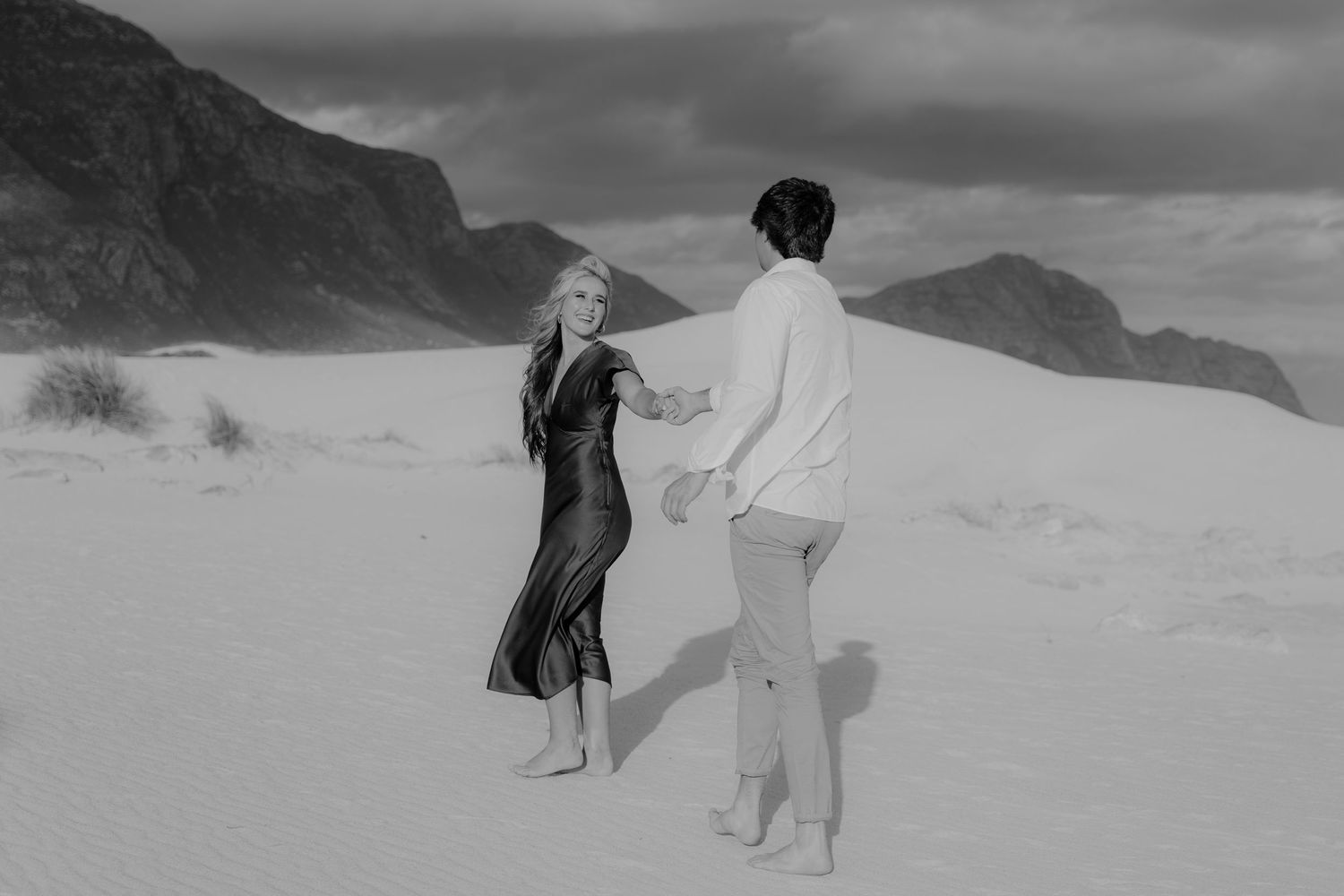 Black and white photo of a couple holding hands and dancing barefoot on white sand dunes with mountains in background.