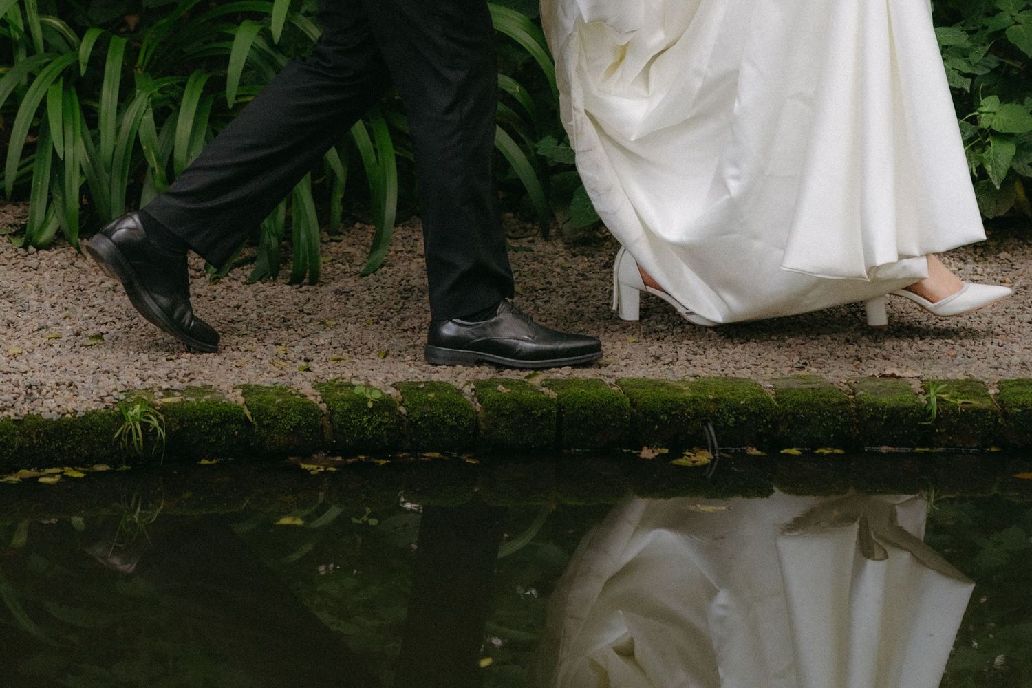 Wedding shoes and dress hem reflected in still water beside a garden path.