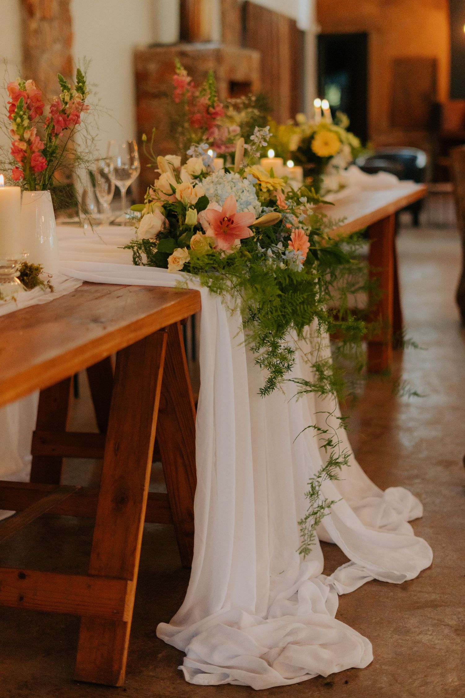 Elegant wedding reception table decorated with flowing white fabric and colorful floral arrangements on wooden benches.