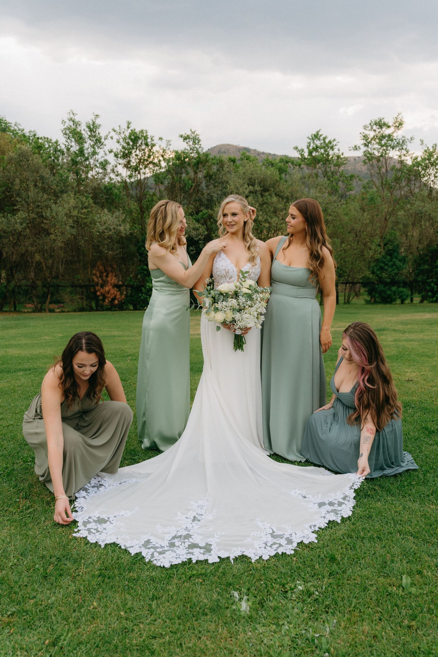 Bridesmaids in sage green dresses pose with the bride's lace train on green grass.