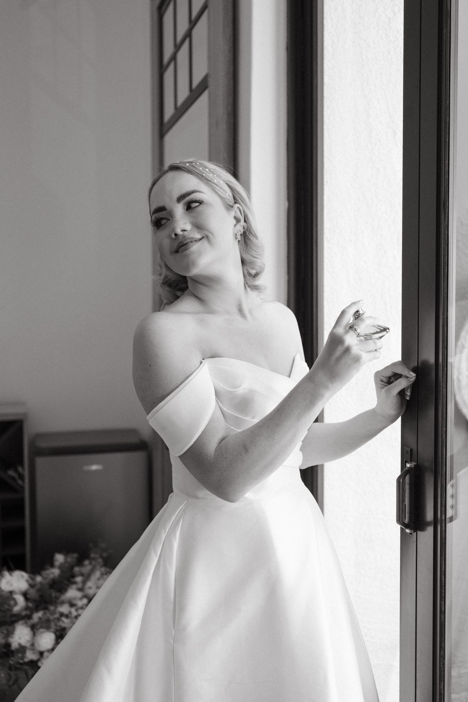 Black and white image of someone in a classic off-shoulder wedding gown standing by a window.