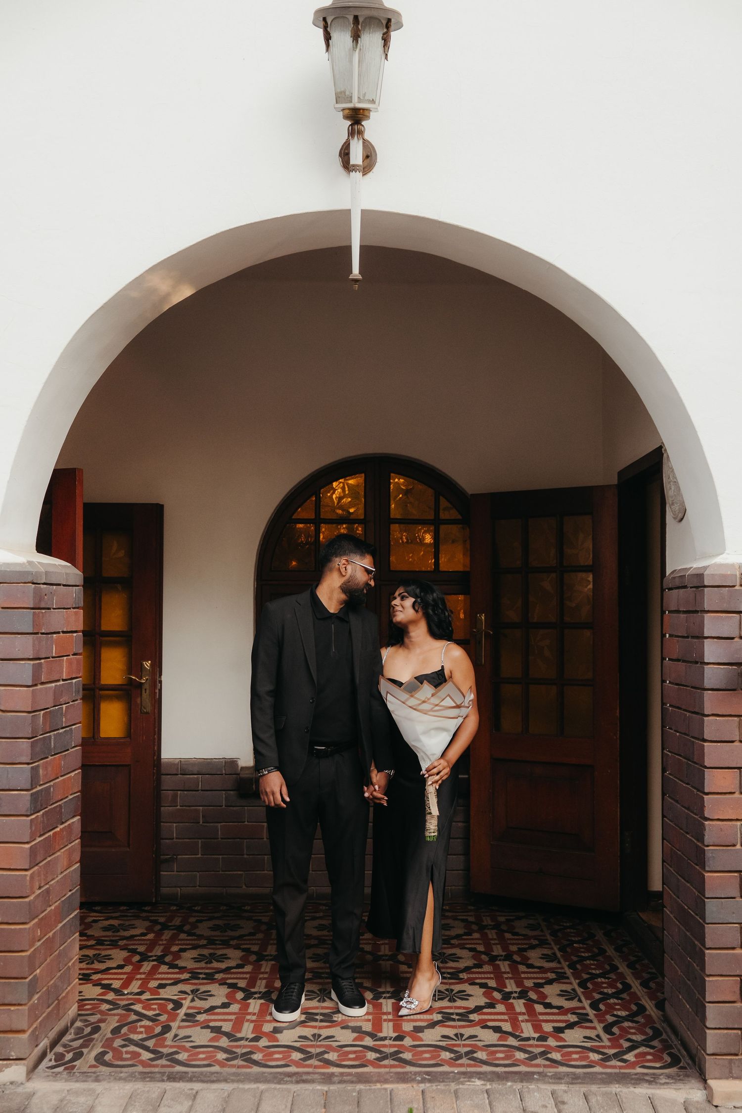 Elegant couple in formal attire standing under a white arched doorway with brick columns and ornate tiled floor.