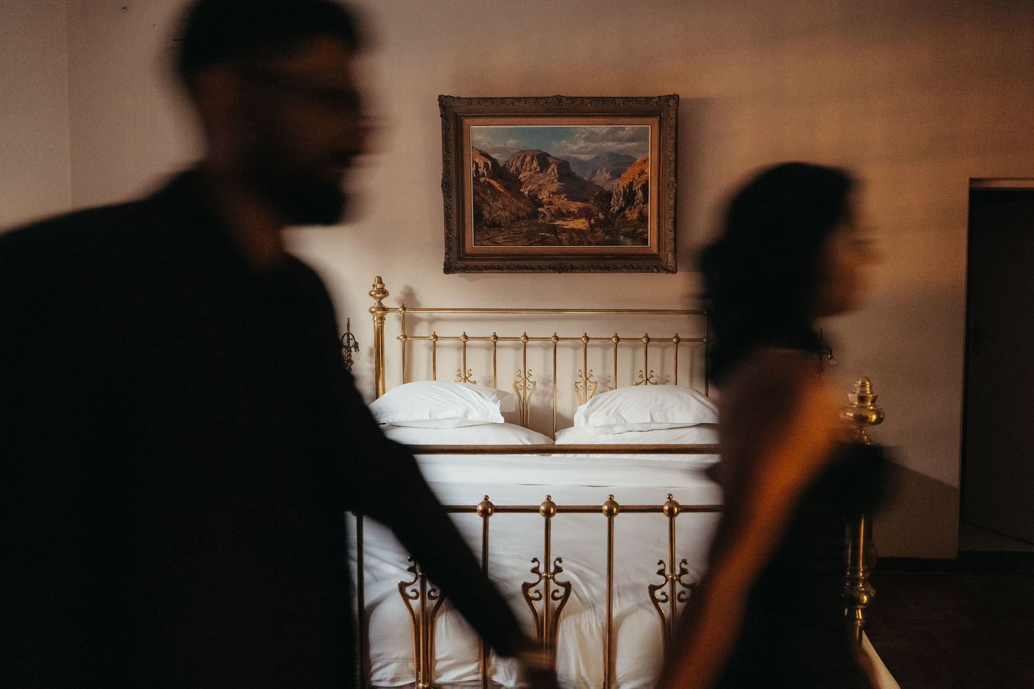 Couple holding hands walking past a vintage brass bed with white pillows and landscape painting in romantic hotel room.