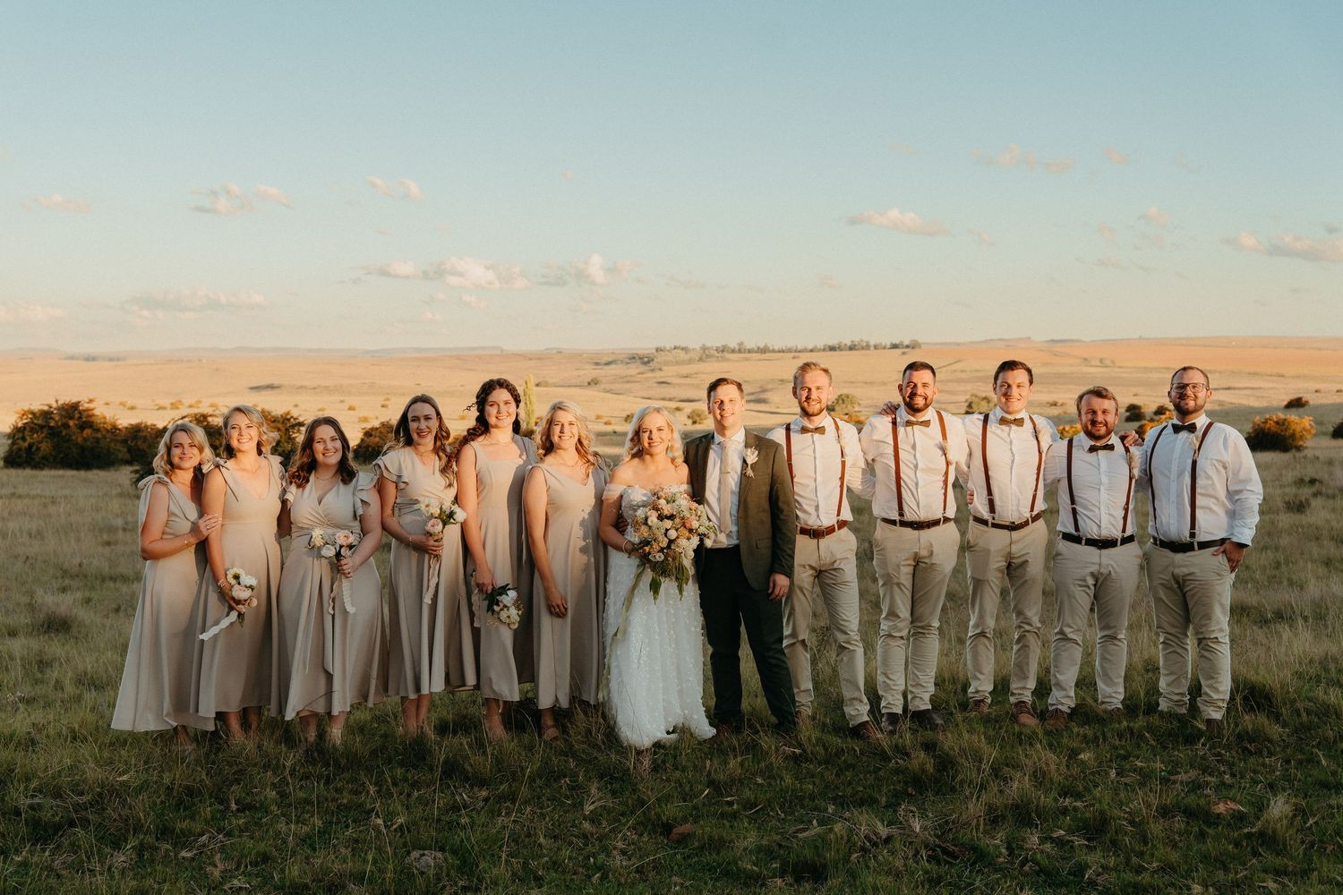Wedding party poses in golden field at sunset, bridesmaids in blush gowns and groomsmen in tan suits with suspenders.