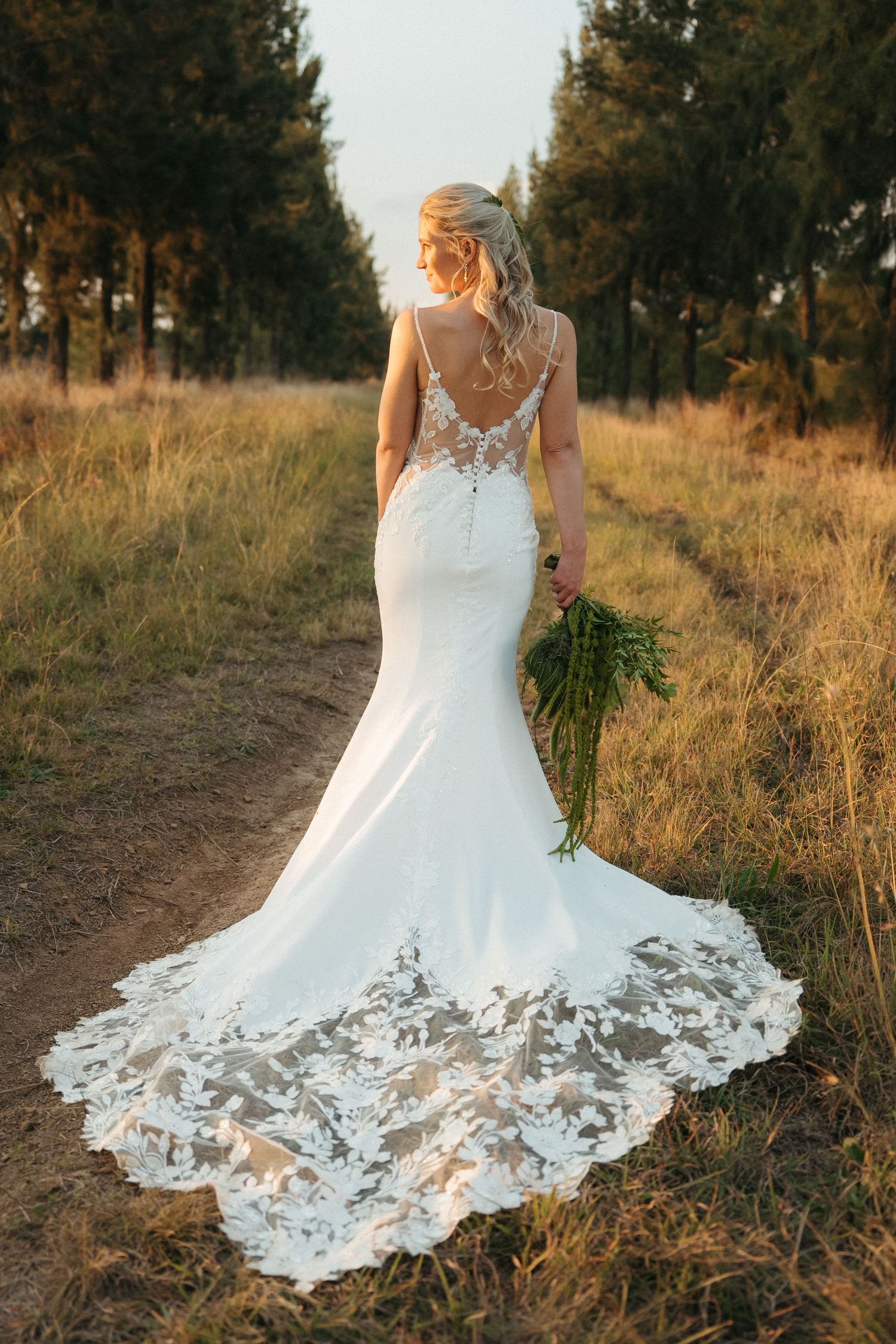 Bride in a fitted lace mermaid wedding gown with a floral train walks a golden field path holding a green bouquet.