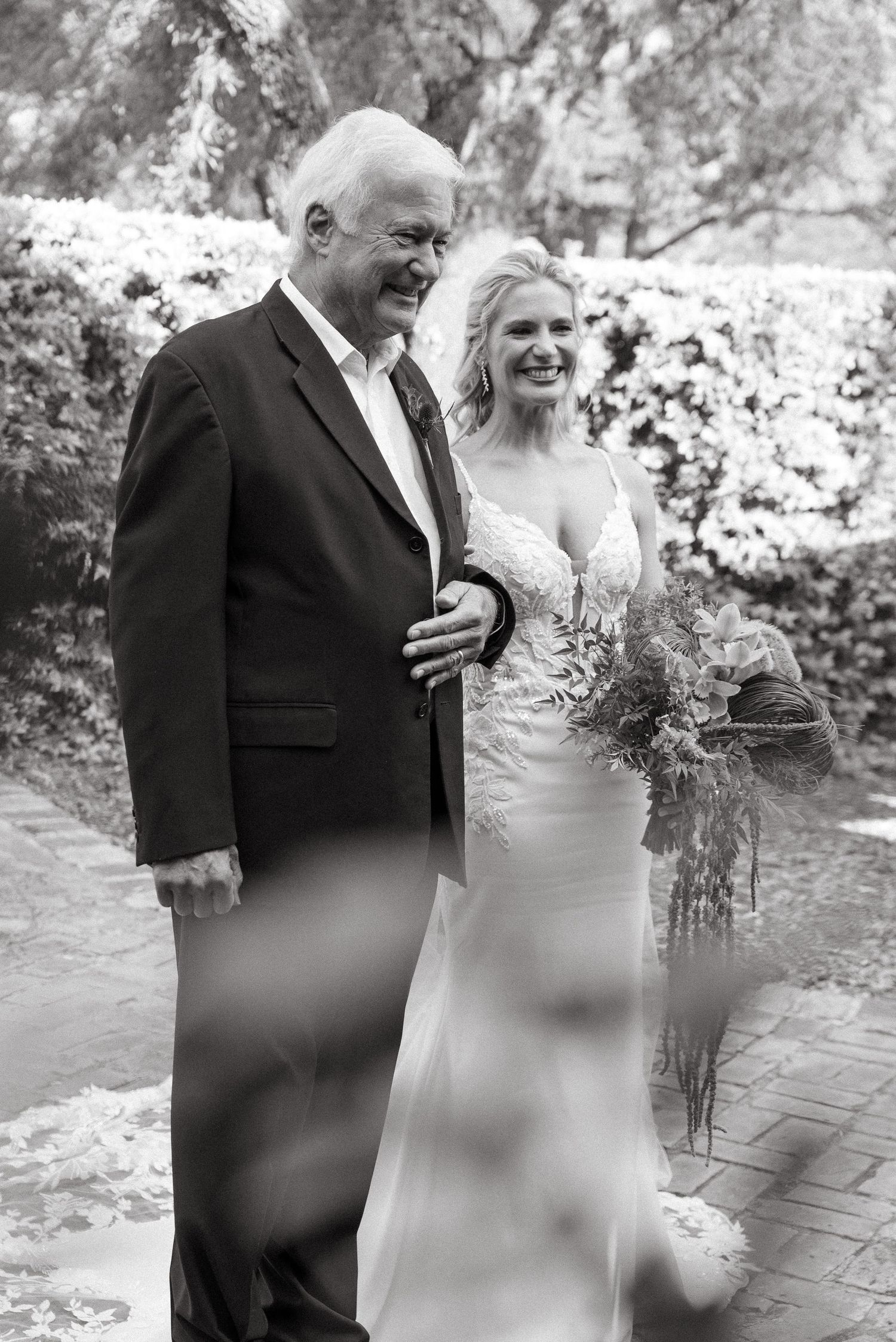Black and white photo of a smiling bride in lace gown holding bouquet, walking with older man in suit.