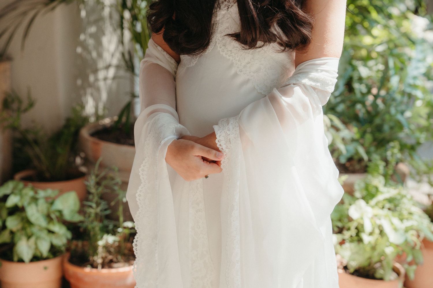 Bride in a white strapless wedding gown with sheer long sleeves poses among potted plants in a garden setting.