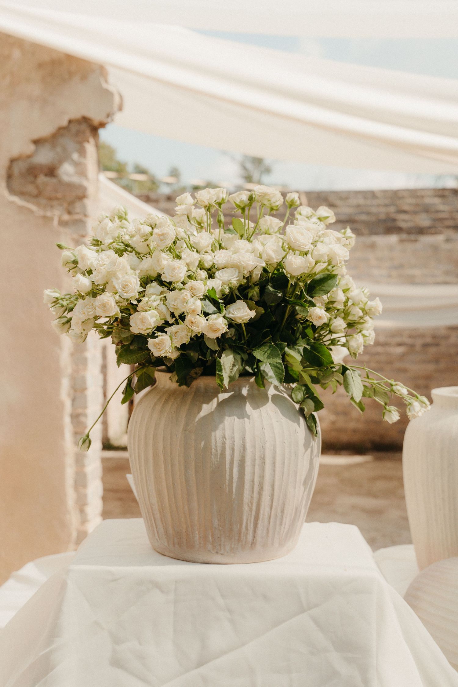 White floral arrangement in a ribbed ceramic vase on a white draped table at an outdoor wedding venue.