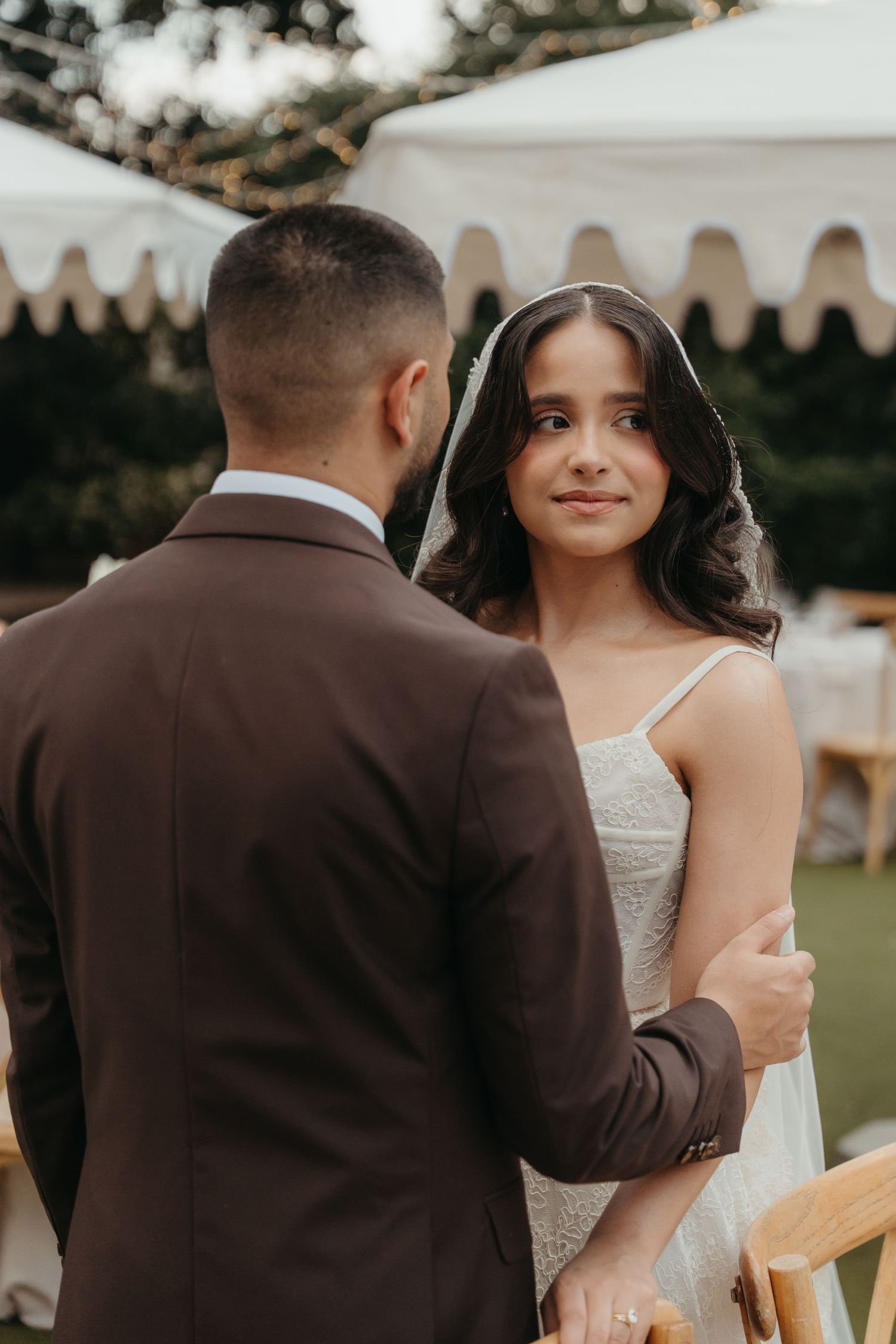 Bride in lace gown and veil smiles at groom in brown suit during outdoor wedding ceremony.