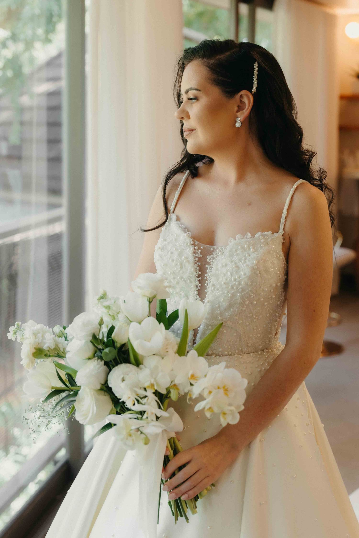 Bride in beaded wedding gown holds white floral bouquet, gazing out a window with soft natural light.