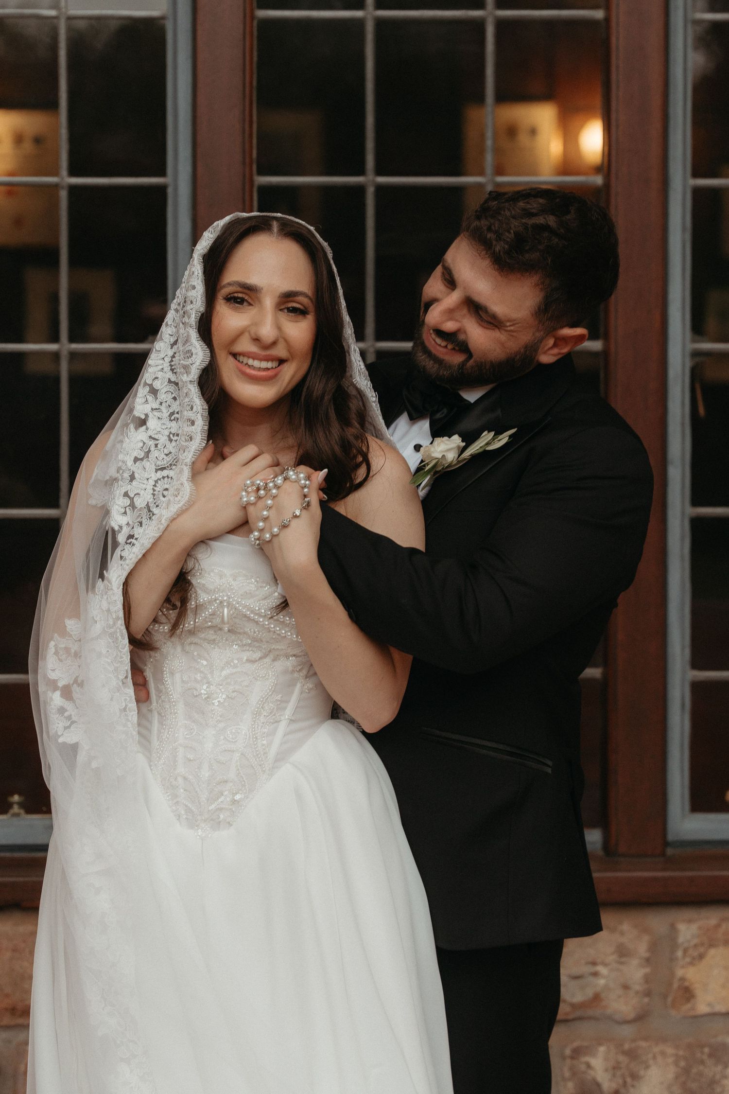 Bride in lace veil and white gown smiles as groom in black tuxedo clasps pearl necklace around her neck.