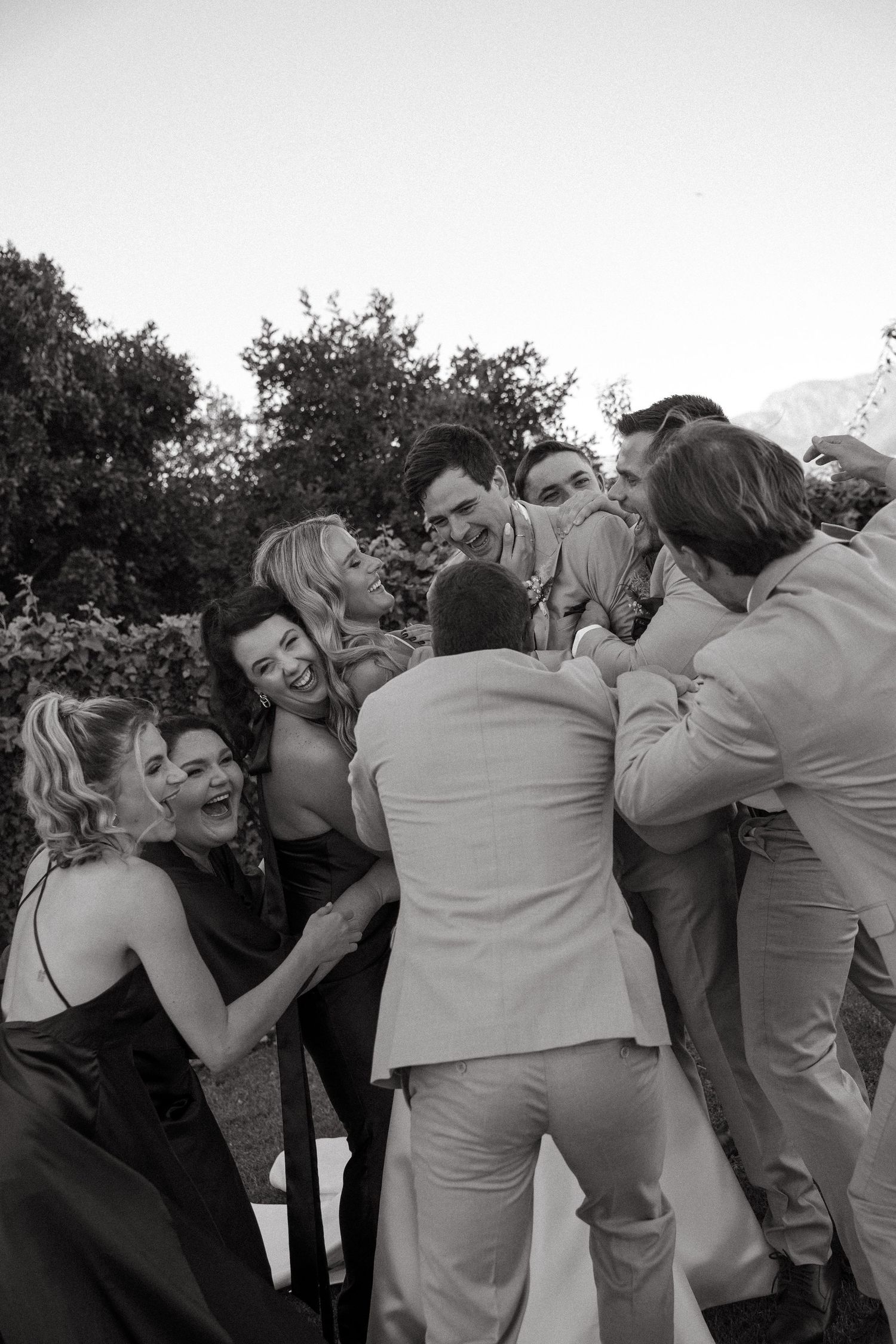 Black and white photo of laughing wedding guests in formal attire huddled together in a joyful group embrace outdoors, with trees behind.