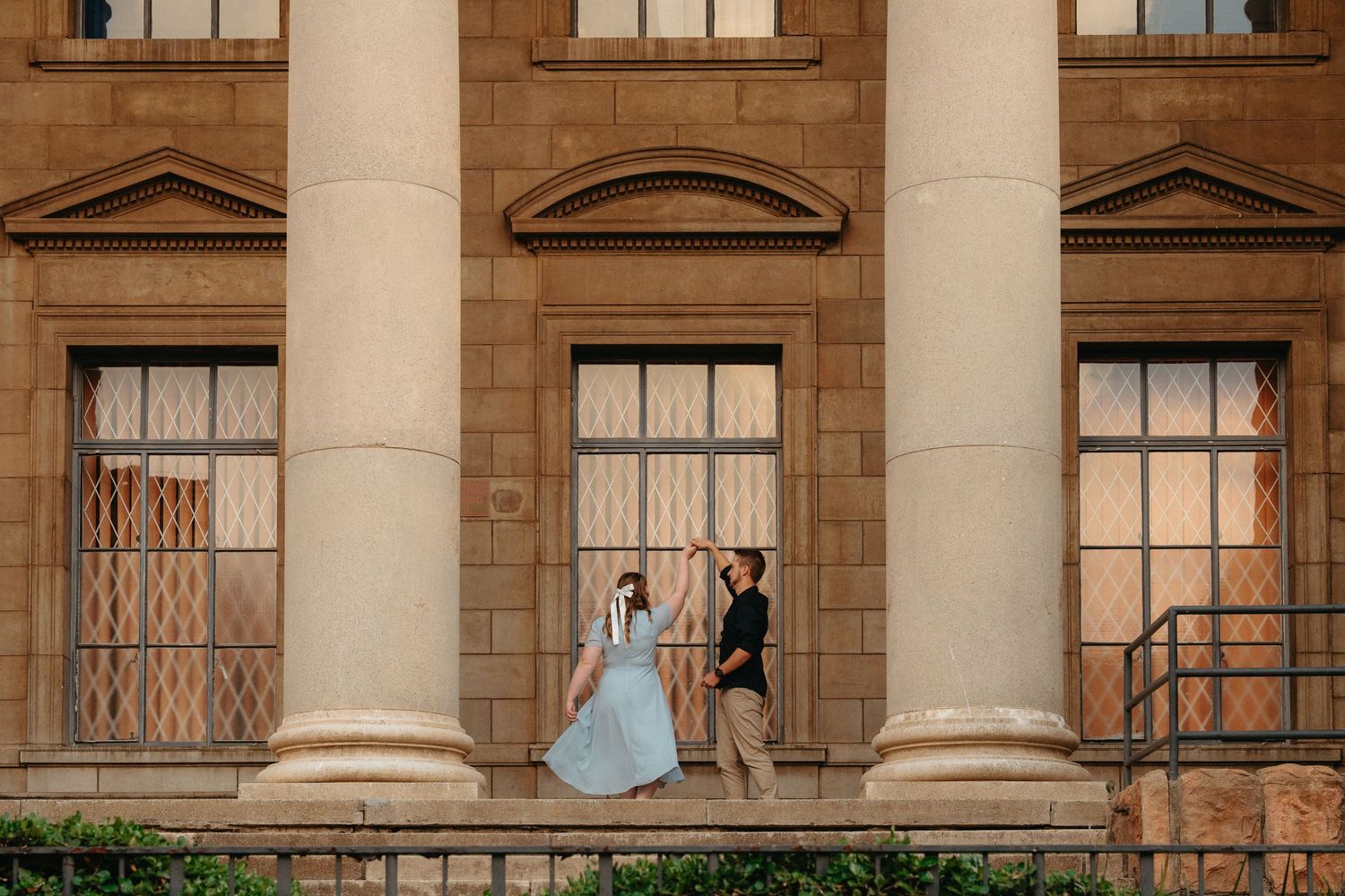 Couple walking hand in hand along grand columned WITS building facade during romantic engagement photo session.