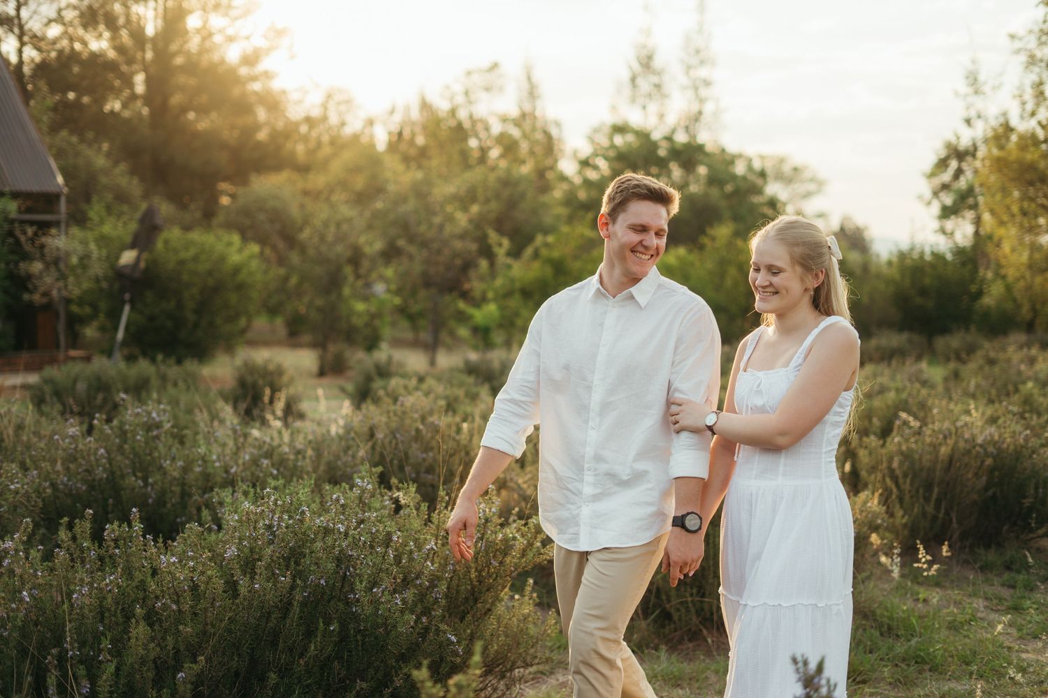 A couple takes an evening stroll through a sunlit meadow wearing white and beige attire at golden hour.