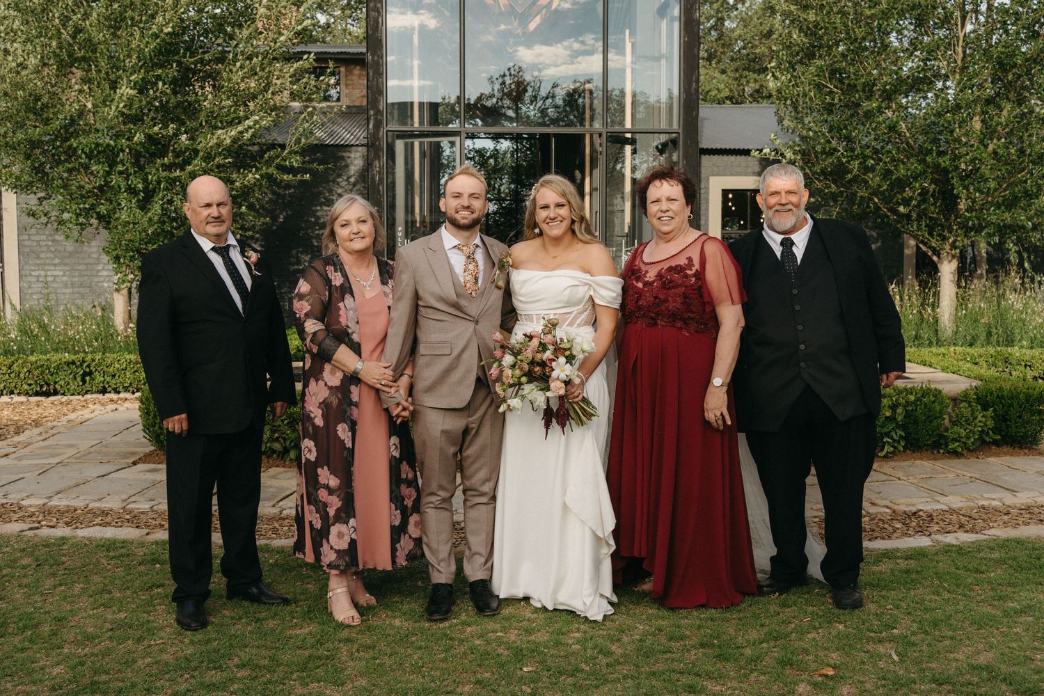 A wedding party poses together outdoors in formal attire against a garden backdrop on a sunny day.