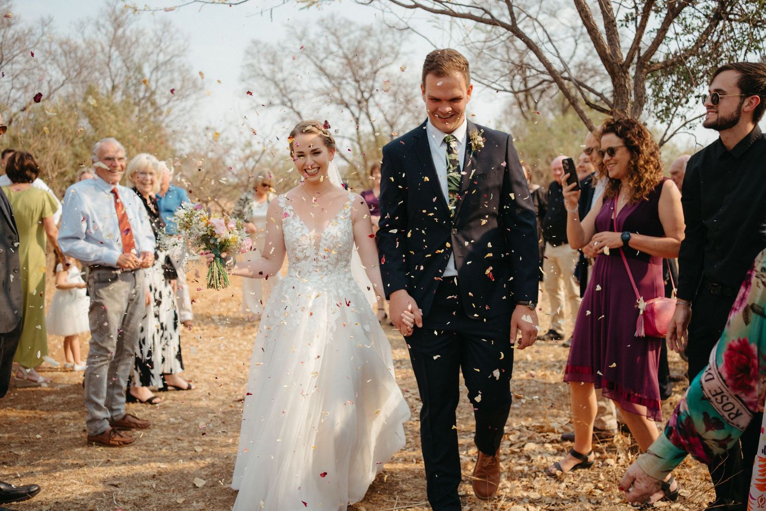 Newlyweds walk through shower of confetti on outdoor path while wedding guests celebrate.