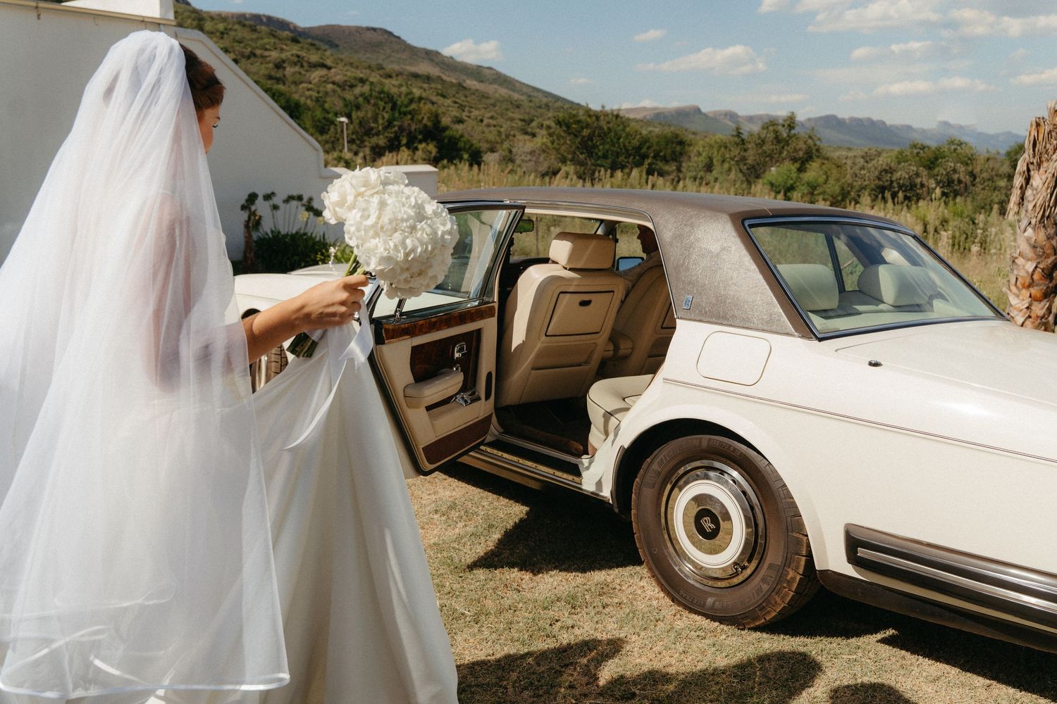 A bride in a flowing white dress walks through a desert landscape with vintage cars and mountains in the background.