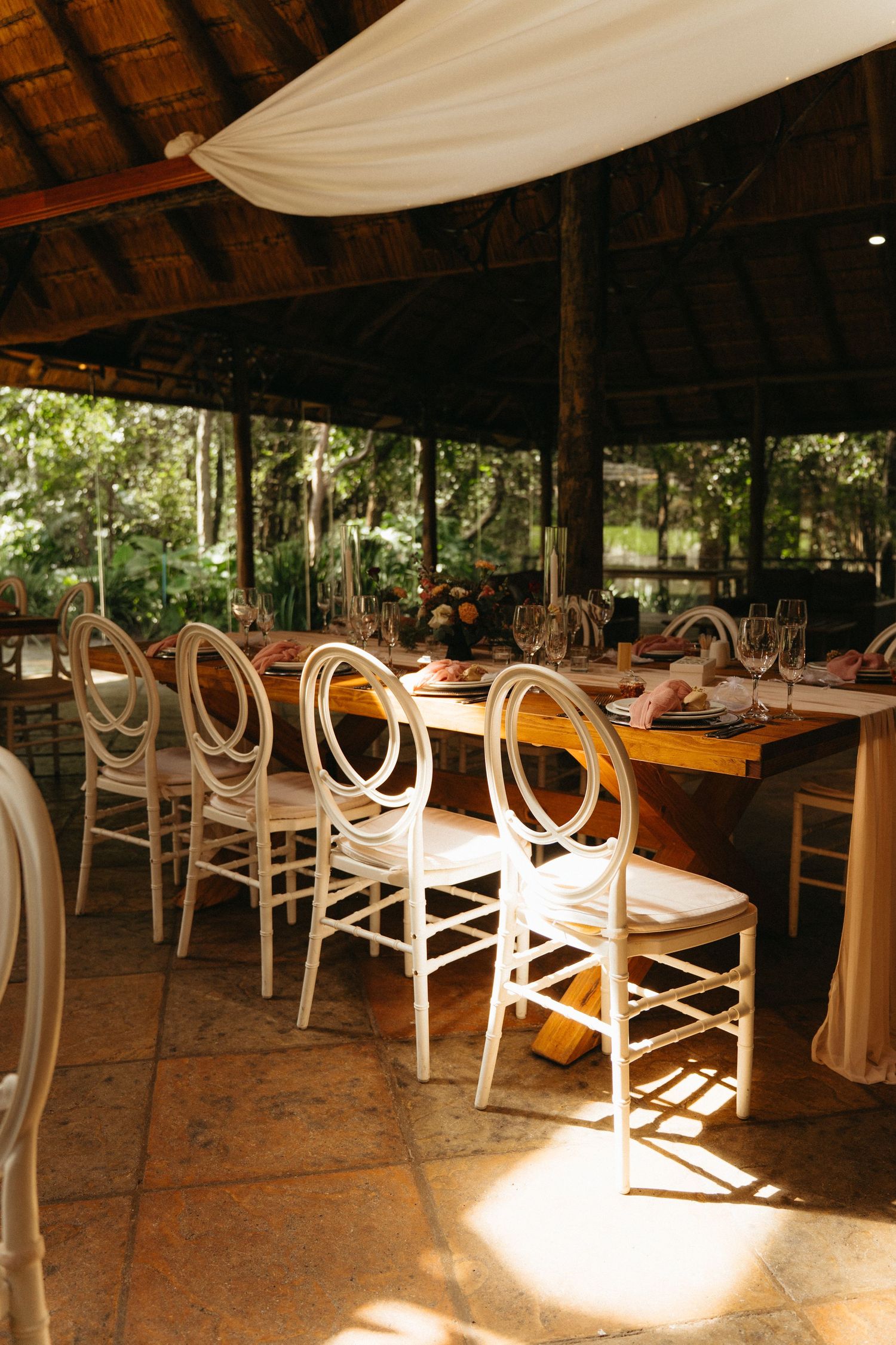 Elegant white phoenix chairs line a rustic outdoor dining setup under a thatched roof pavilion with warm sunlight streaming in.