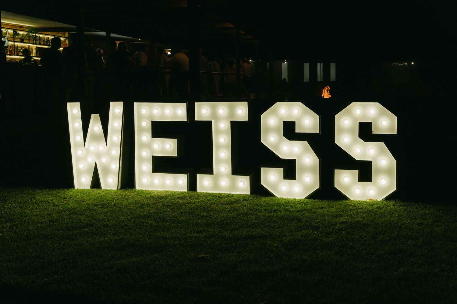 Large illuminated white WEISS letters glowing on dark grass at night create a striking display.