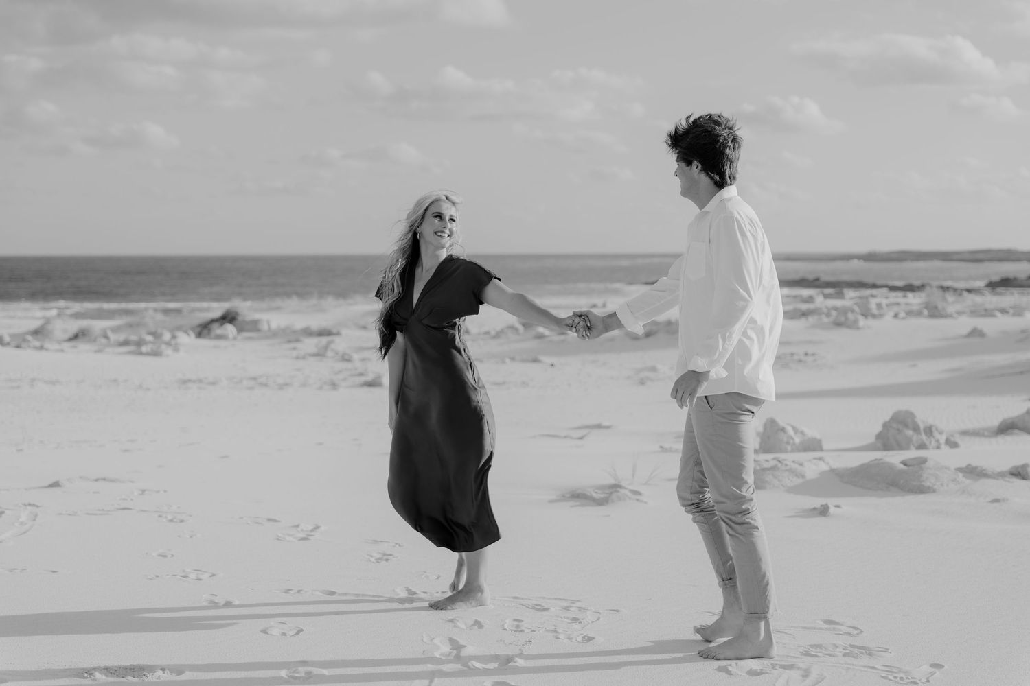 Black and white photo of a couple holding hands on a sandy beach with ocean waves in the background.